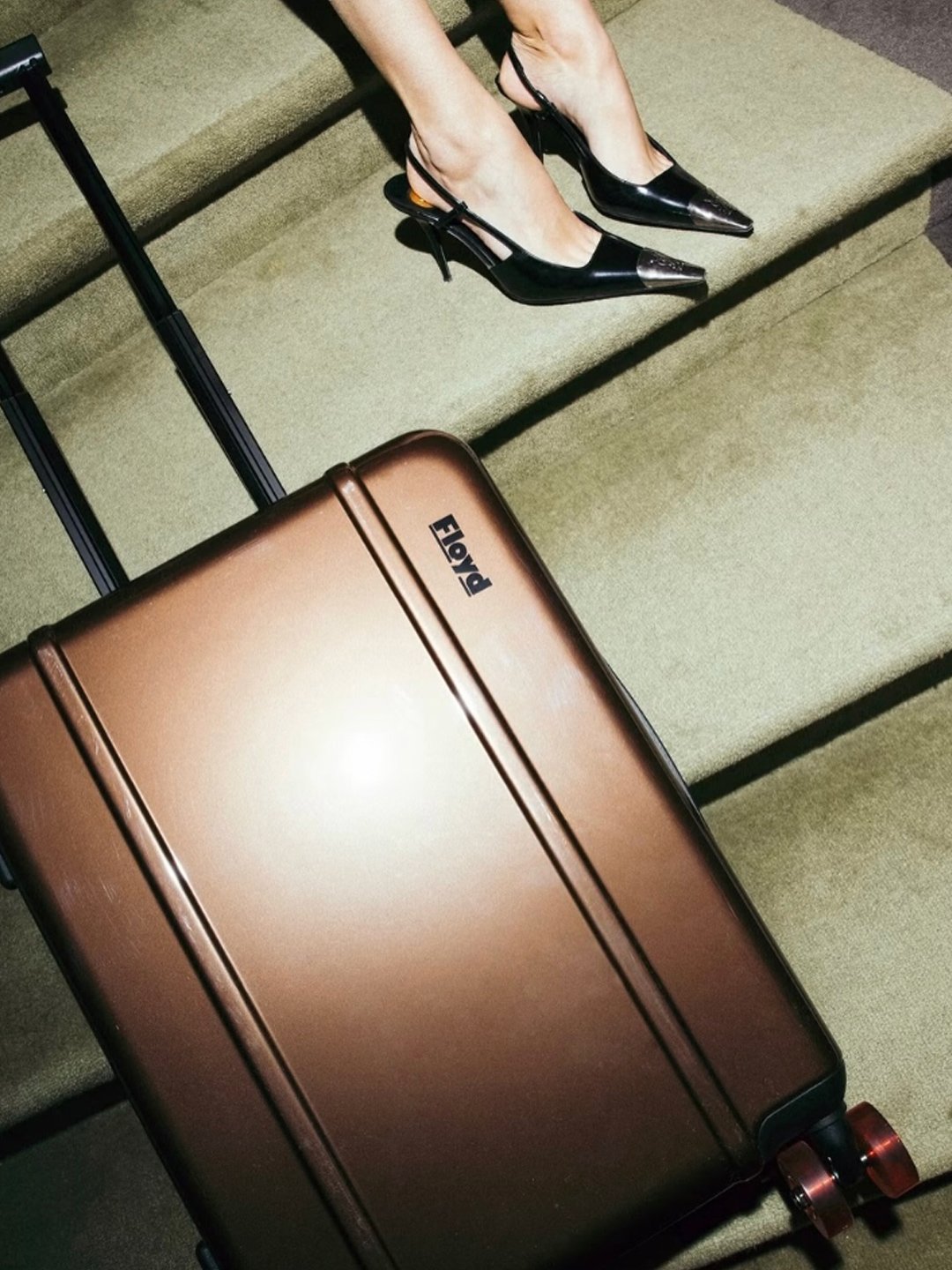 Person sitting on stairs wearing black high-heeled shoes with pointed toes and carrying a tan hard-shell suitcase.