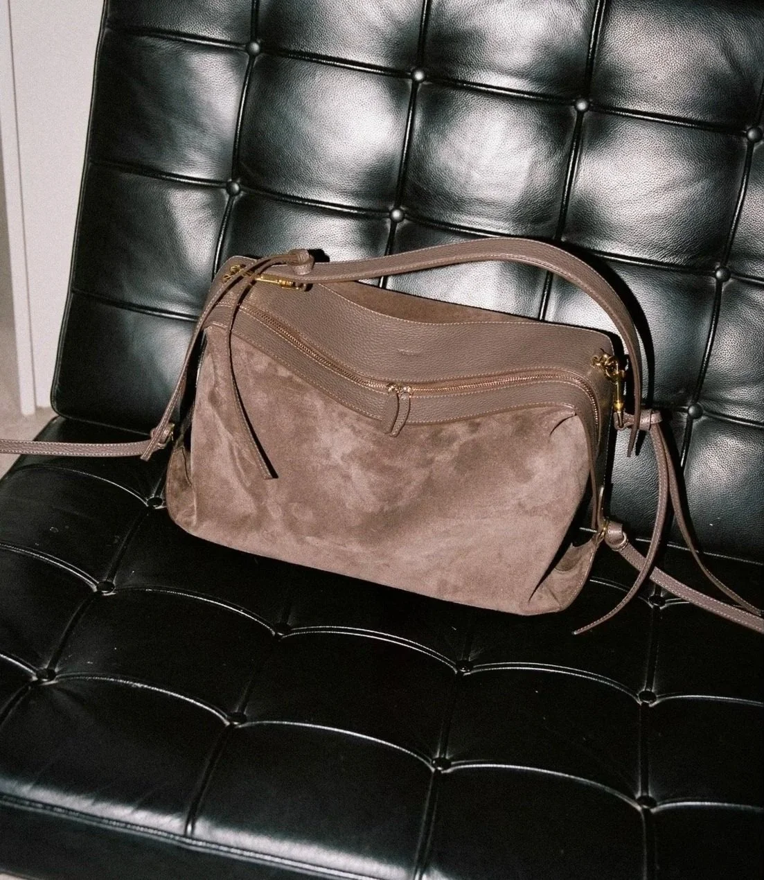 A taupe-colored handbag with gold accents resting on a black tufted leather bench.