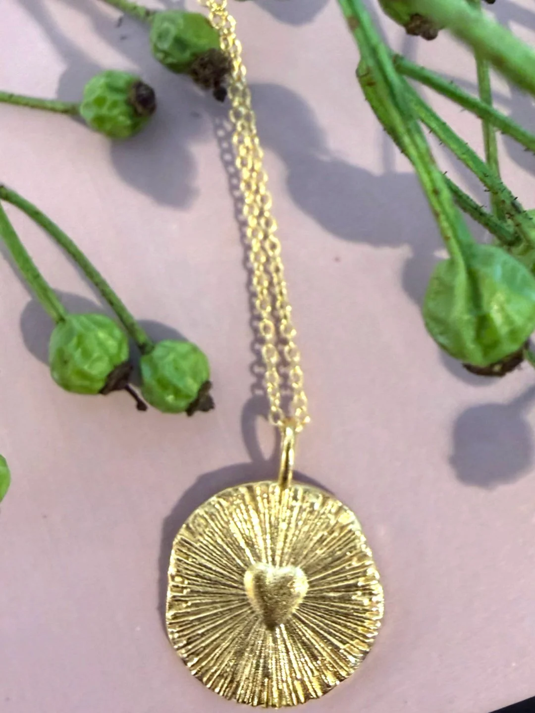 Gold necklace with a circular pendant featuring a heart in the center, surrounded by radiating lines, with green berries and small green leaves around it.
