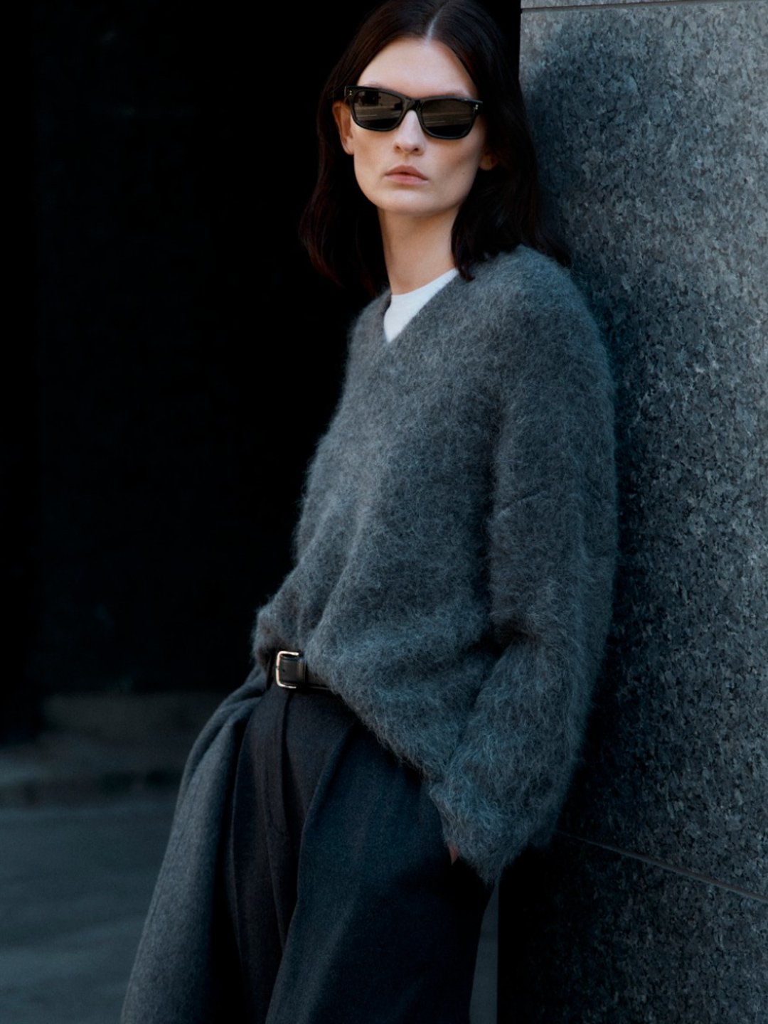 A woman with dark hair wearing sunglasses, a gray sweater, and dark pants, leaning against a textured stone wall.