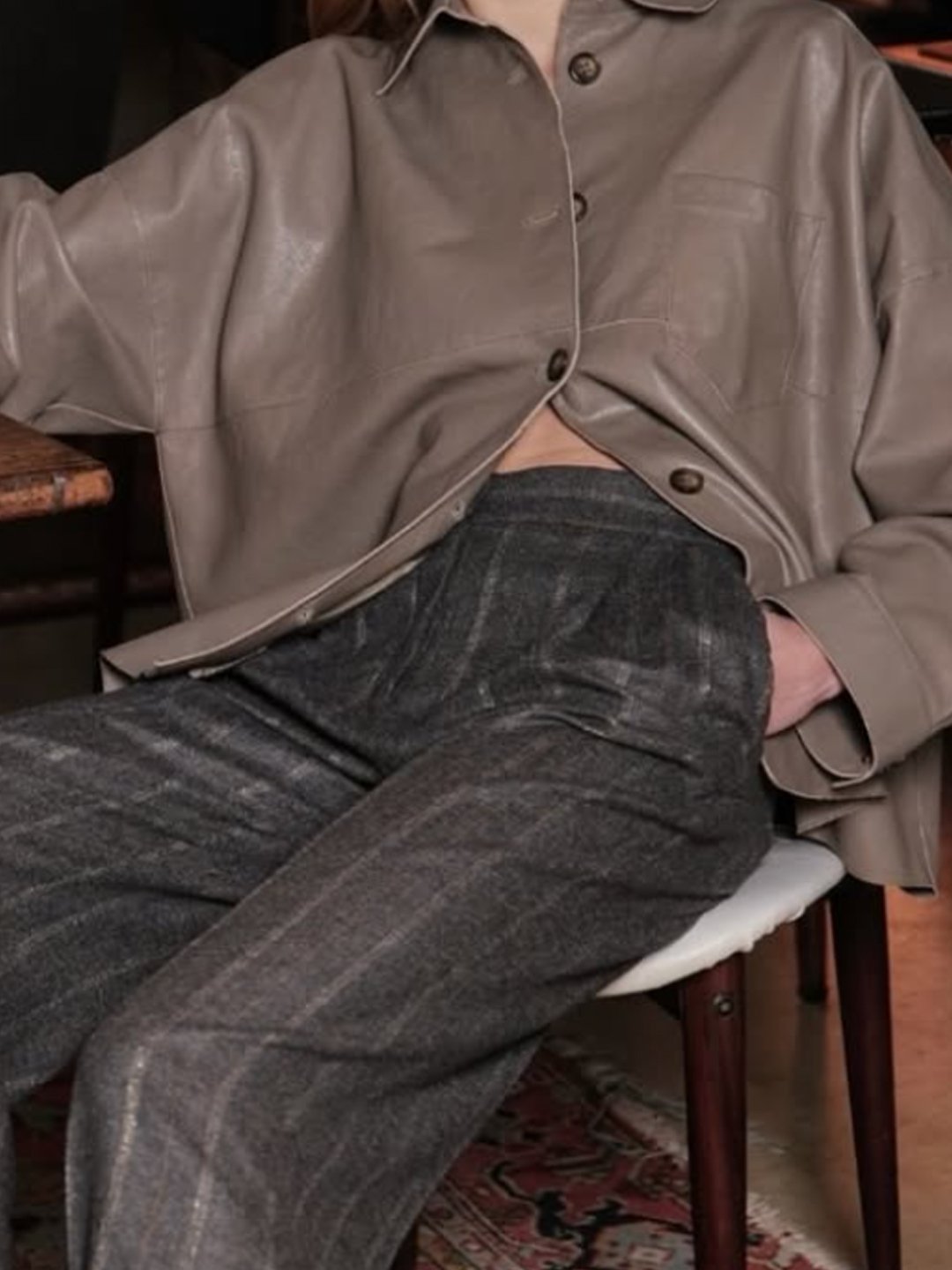 A person sitting on a chair wearing a taupe-colored leather jacket and gray plaid pants, with one hand in their pocket.