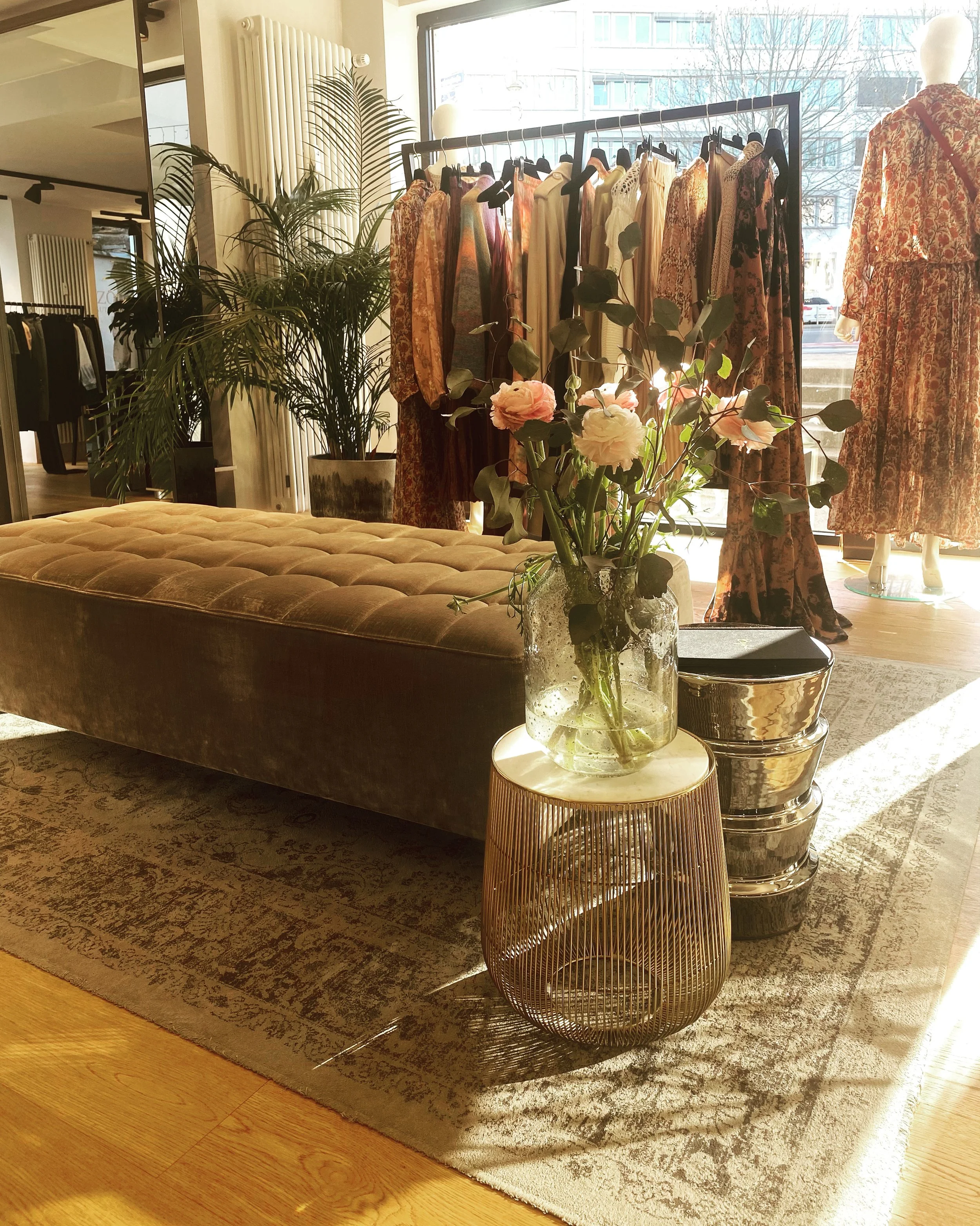 Fashion boutique interior with a clothing rack displaying dresses and garments, a mannequin by the window, a large potted plant, a plush beige ottoman, a glass vase with pink and white flowers, and metallic side tables, lit by natural sunlight.
