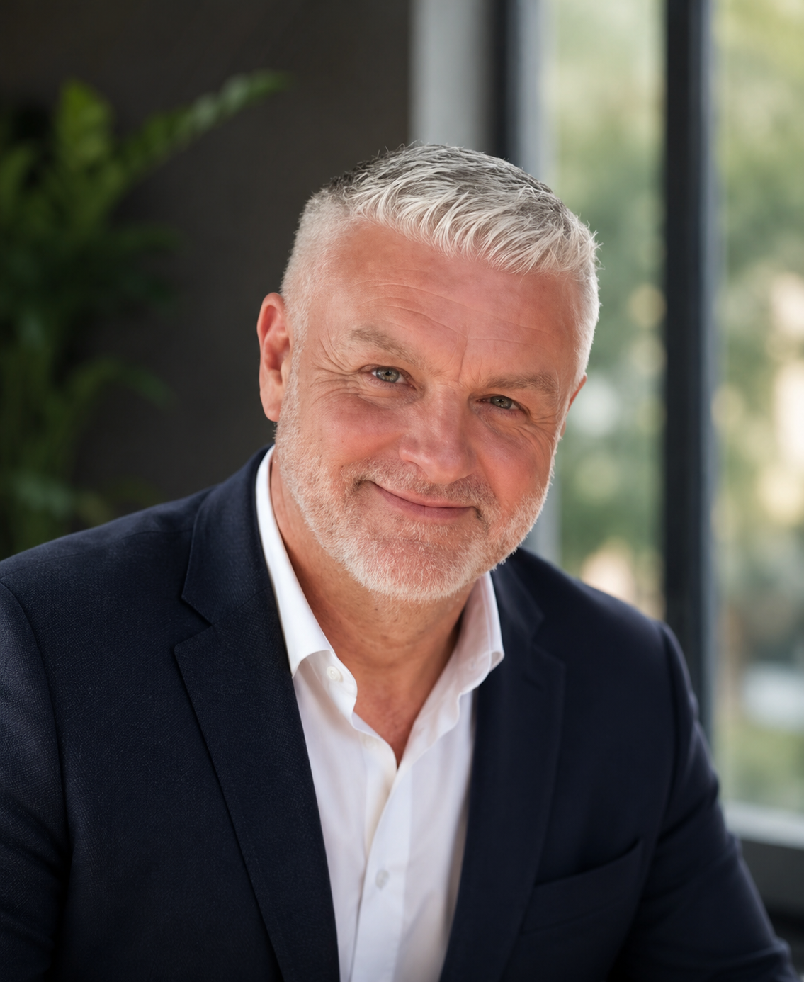 A middle-aged man with gray hair and a beard, wearing a dark suit and white shirt, smiling at the camera in a modern office setting with large windows and greenery outside.