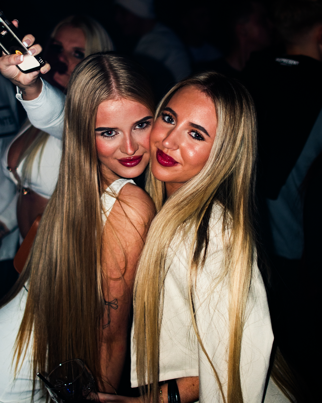 Two women with long blonde hair and makeup, smiling and posing for a photo at a social event or party.