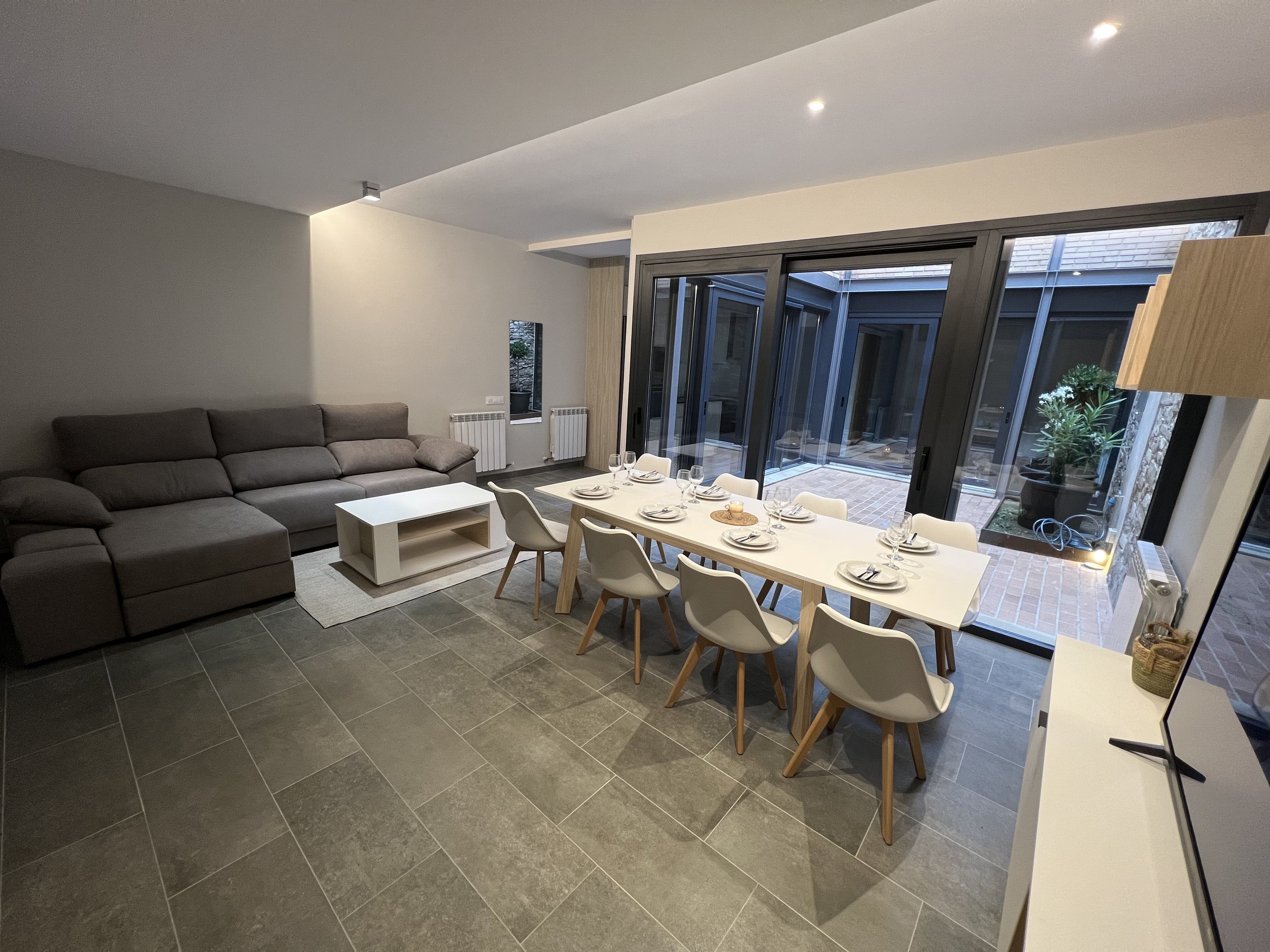 Imagen de una sala moderna con sofá gris, mesa pequeña y comedor con sillas blancas, ventanas grandes que permiten entrada de luz natural y vista a un patio.