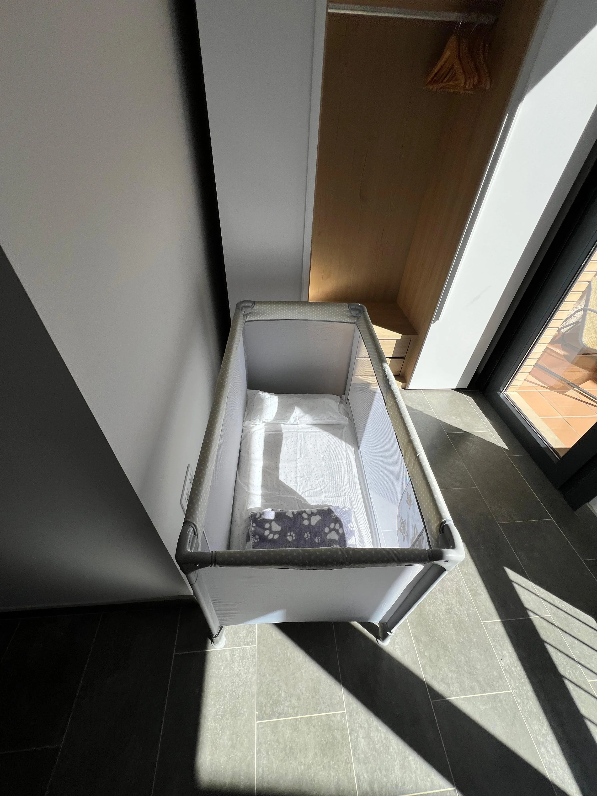 Cuna de bebé en una habitación con luz natural, cerca de una ventana con vista a un balcón. La habitación tiene paredes blancas, piso de baldosas y muebles de madera.