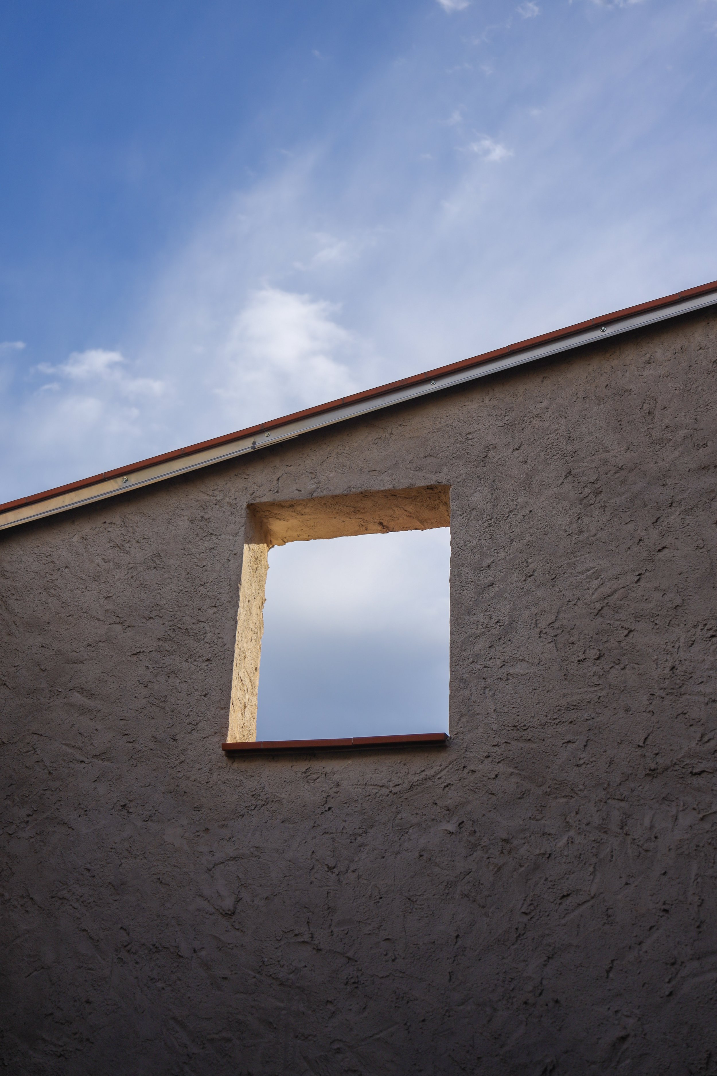 Vista frontal de una construcción de paredes de color beige con dos ventanas sin cristales, una en la parte superior y otra en la parte inferior, ambas con molduras de color terracota. El cielo es parcialmente visible, con nubes dispersas en un fondo azul.