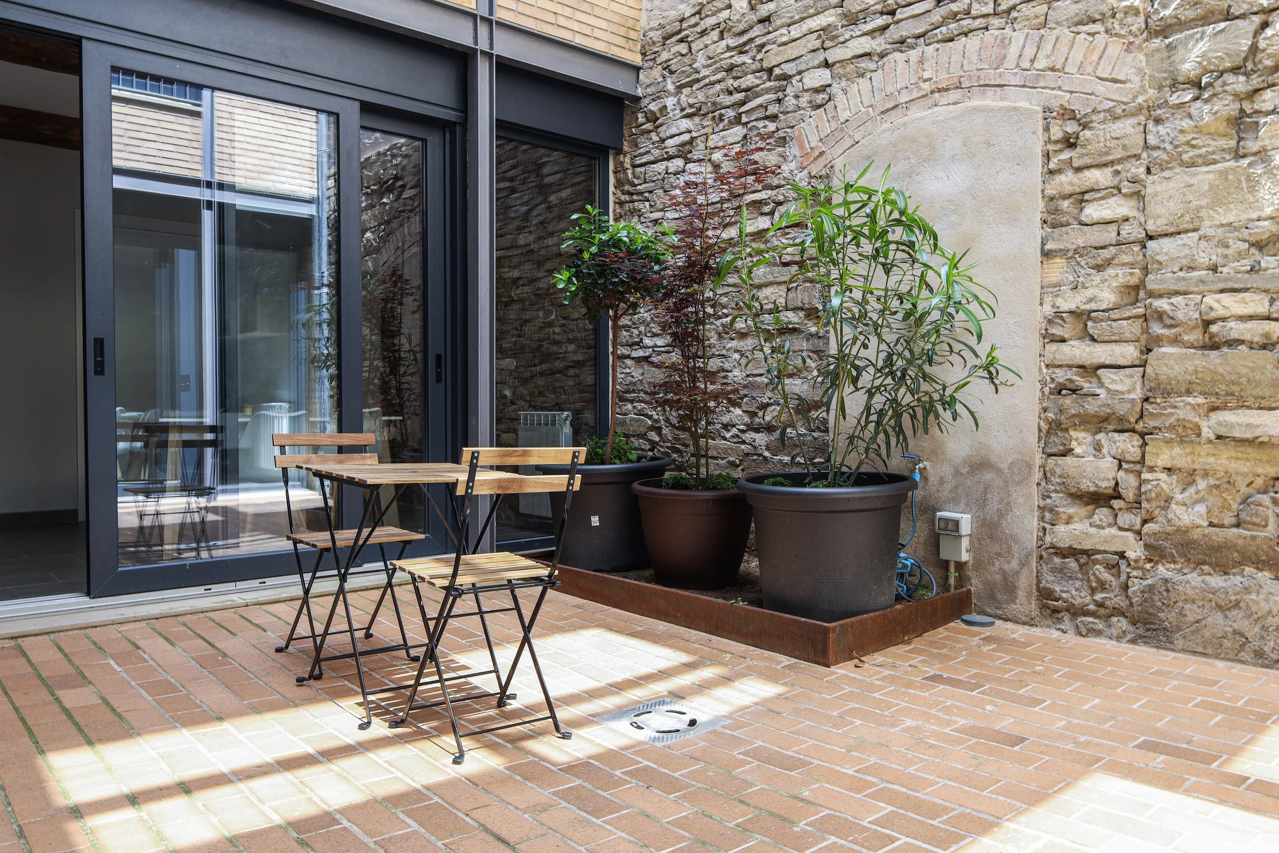 Área de patio o terraza con piso de ladrillo, mesa y sillas de madera, plantas en macetas grandes y muro de piedra y ladrillo.