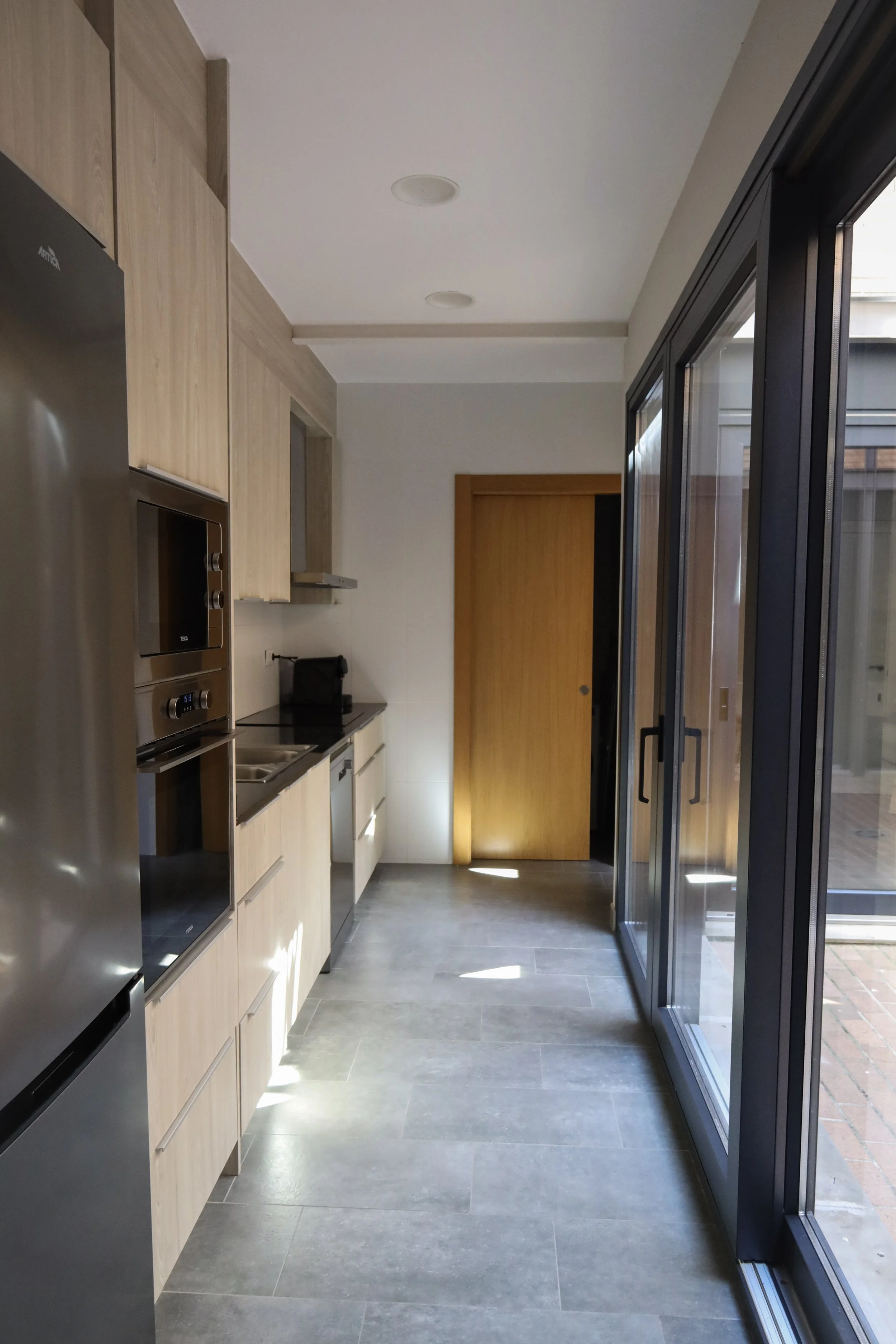 Cocina moderna con gabinetes de madera clara, electrodomésticos de acero inoxidable, puertas corredizas de vidrio, suelo de baldosas grises y ventana grande que deja entrar luz natural.