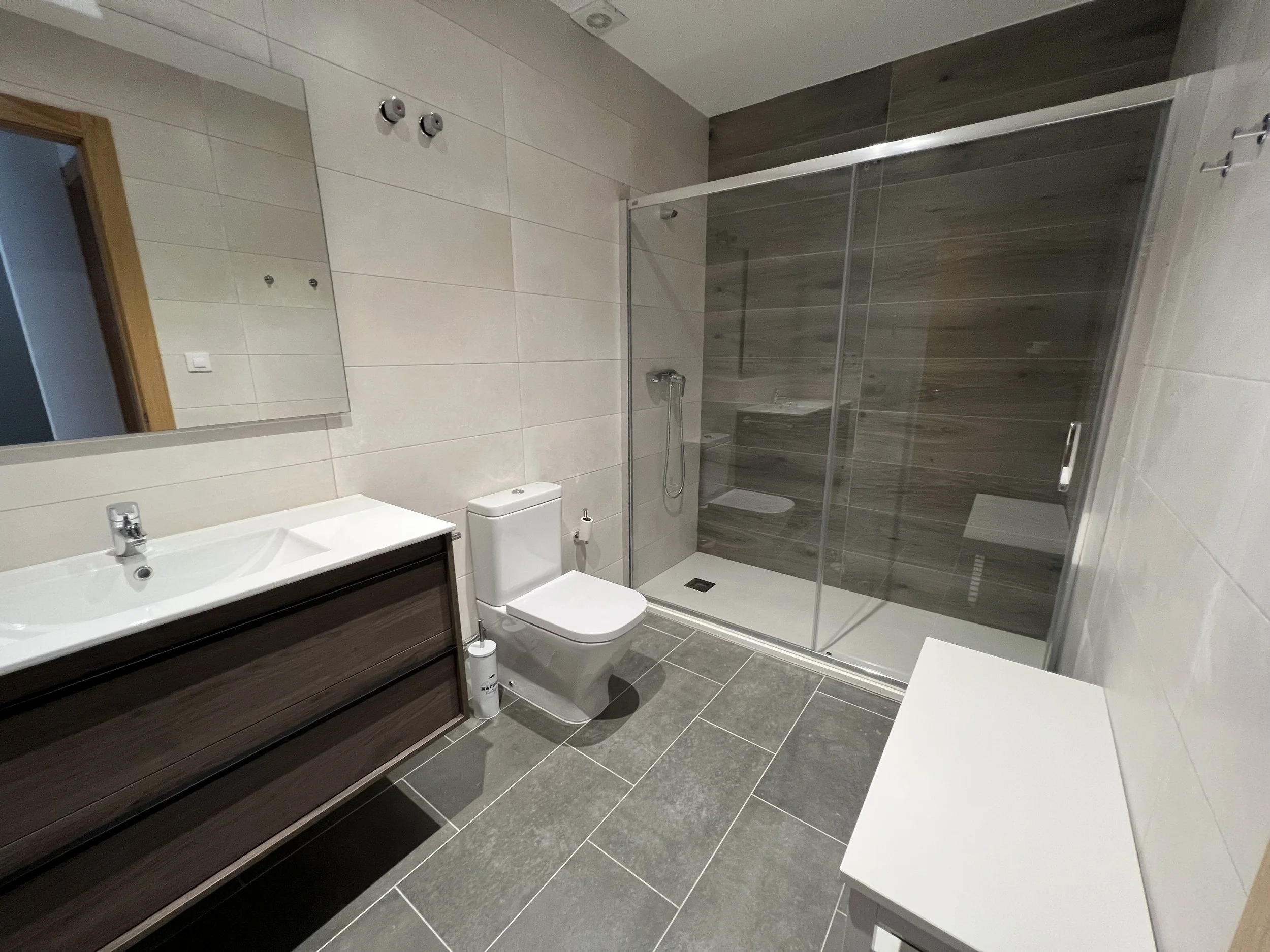 Un baño moderno con paredes y pisos de cerámica gris oscuro y blanco, con una ducha con puertas de cristal, un lavabo con mueble de madera oscura, un espejo grande y un inodoro.