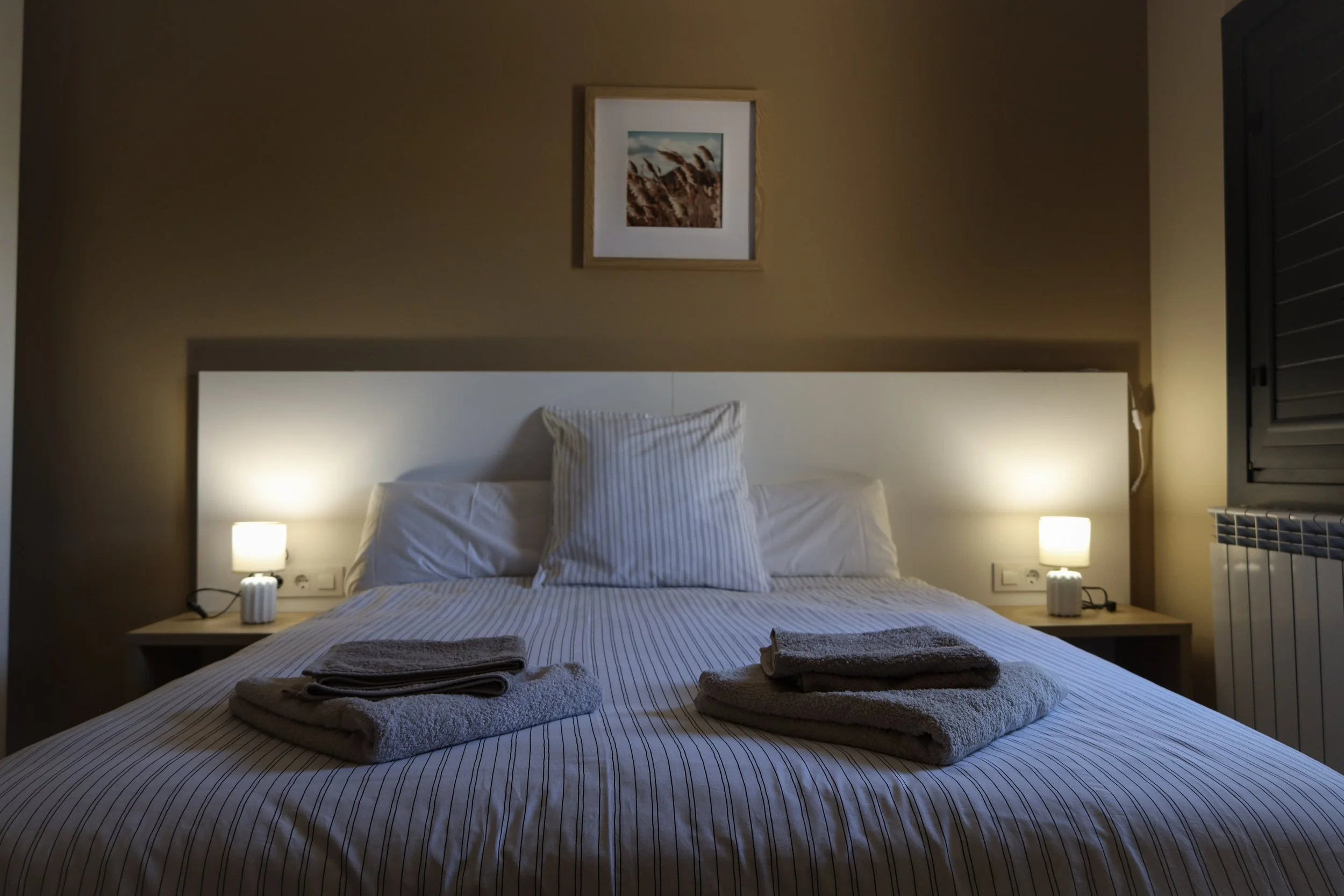 Cama en una habitación moderna con ropa de cama a rayas blancas y grises, dos toallas dobladas en la cama, dos lámparas de noche encendidas a cada lado y una pintura de paisaje en la pared.