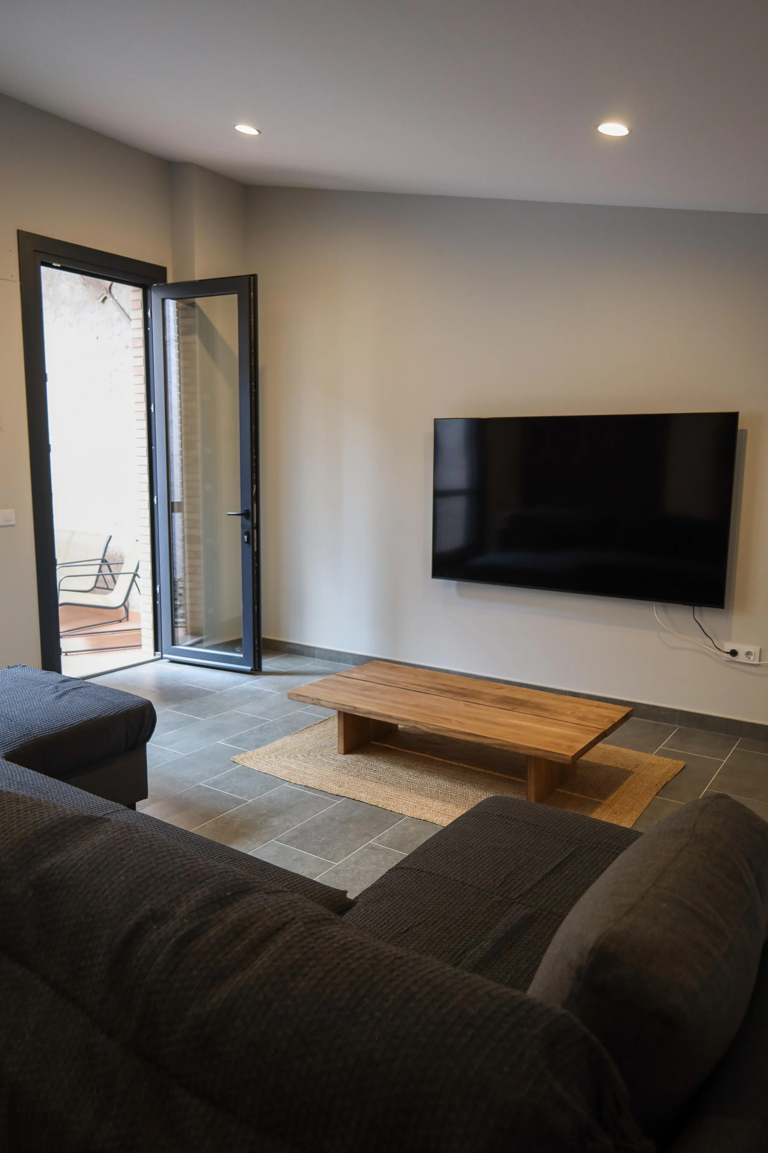 Sala de estar moderna con sofá oscuro, mesa de madera, televisor de pantalla grande en la pared, suelo de baldosas grises, y puertas de vidrio que dan a un balcón con silla de patio.