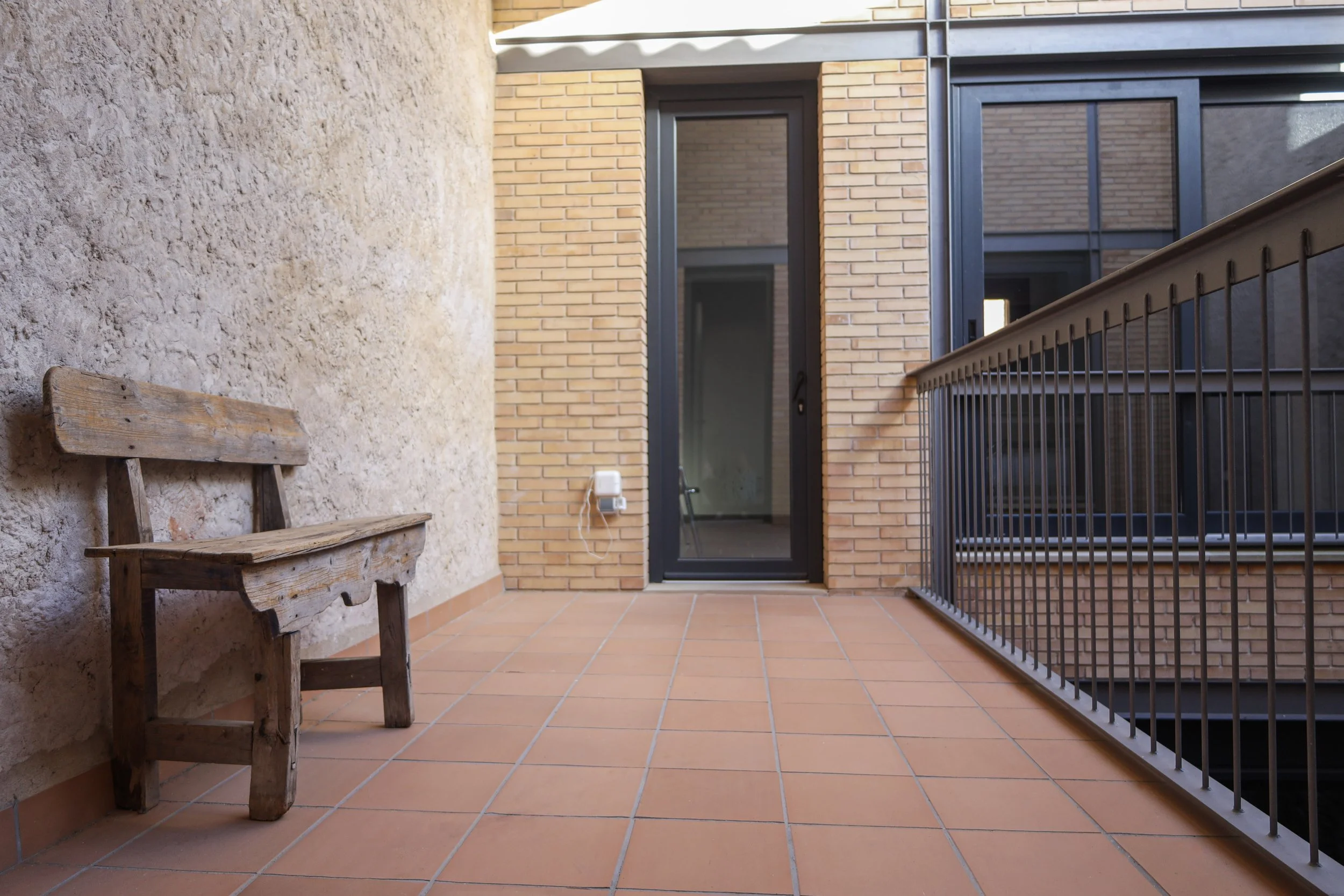 Balcón o terraza urbano con piso de baldosas terracota, muro de piedra y pared de ladrillos, porta de vidrio con marco negro, barandilla de metal, banca de madera y cableado eléctrico en la pared.