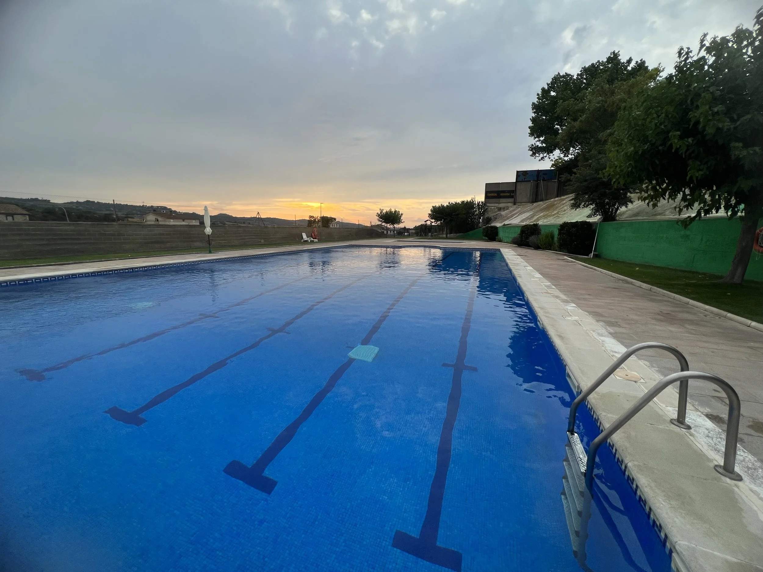 Piscina al atardecer con árboles y construcciones en el fondo, sombra y cielo nublado.