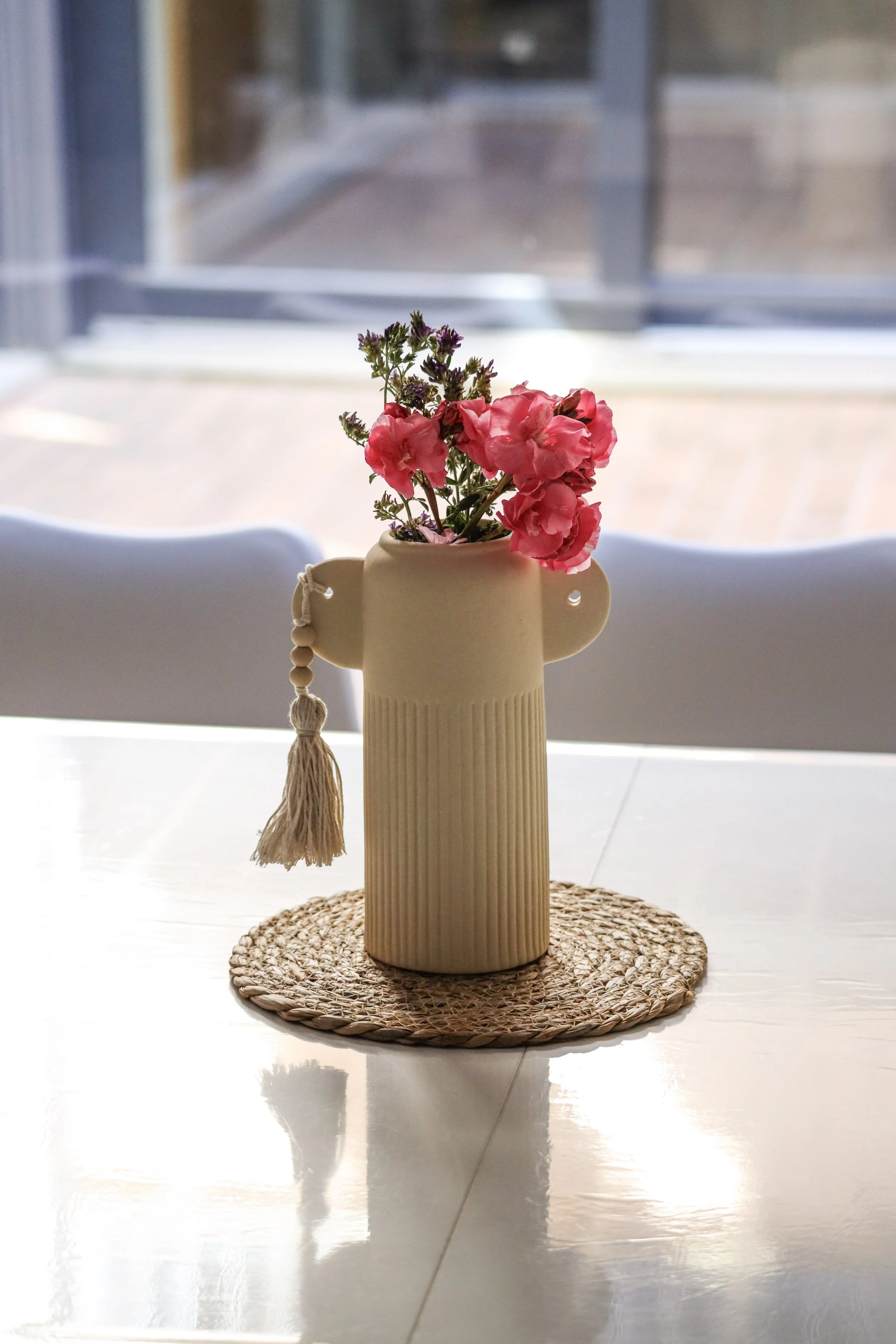 Un jarrón beige con flores rosas y moradas decorativas, colocado sobre un tapete de paja, en una mesa blanca frente a un fondo de ventanales y una pared gris.