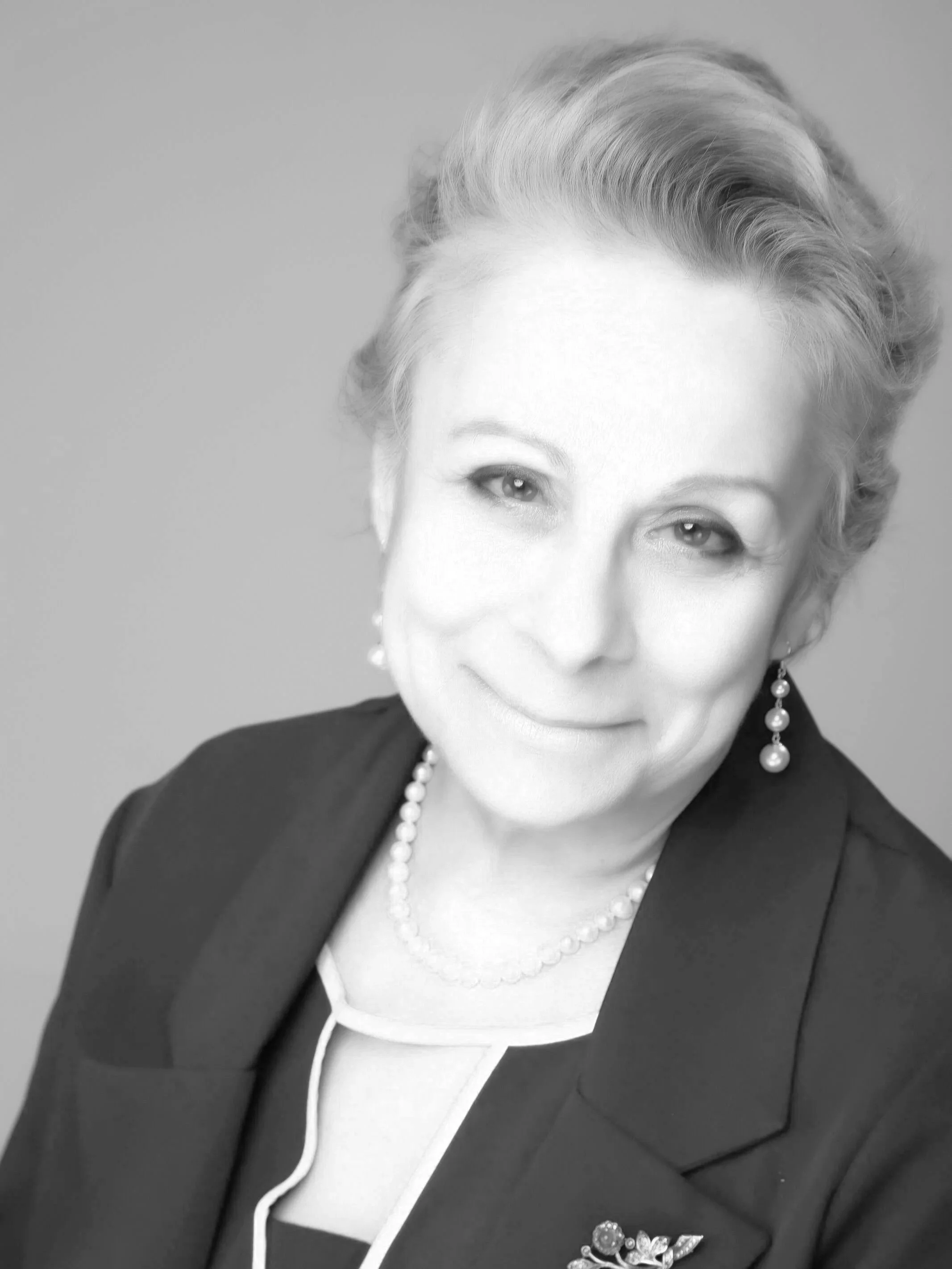 A black-and-white portrait of a woman with styled short hair, wearing earrings, a pearl necklace, and a blazer with a brooch.