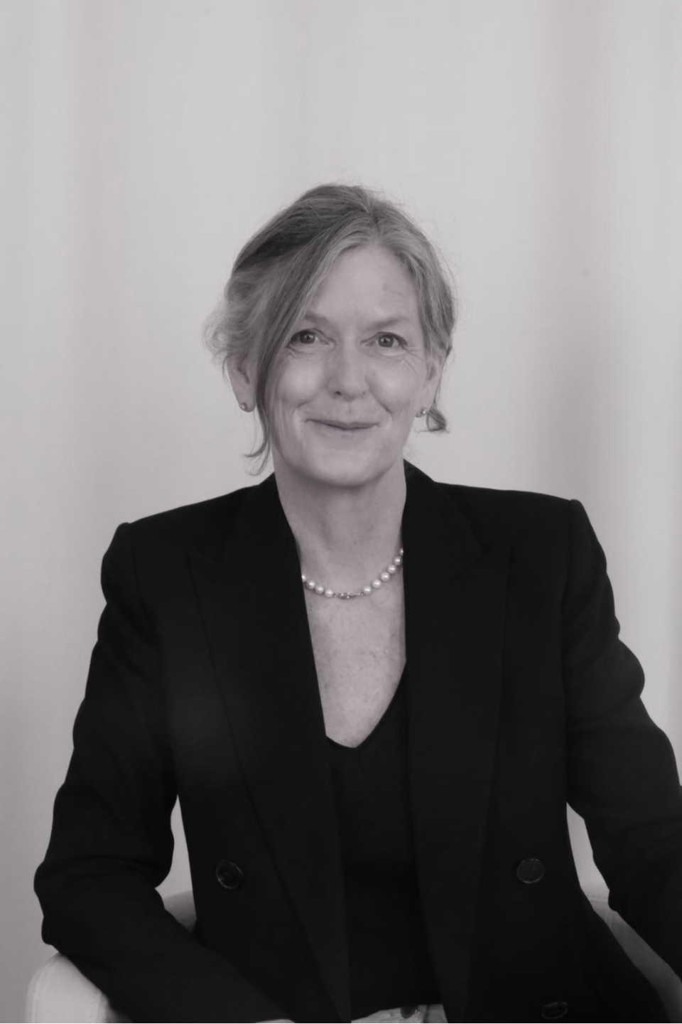 Black-and-white portrait of a woman with short, wavy hair wearing a dark blazer and a pearl necklace, sitting against a plain background.