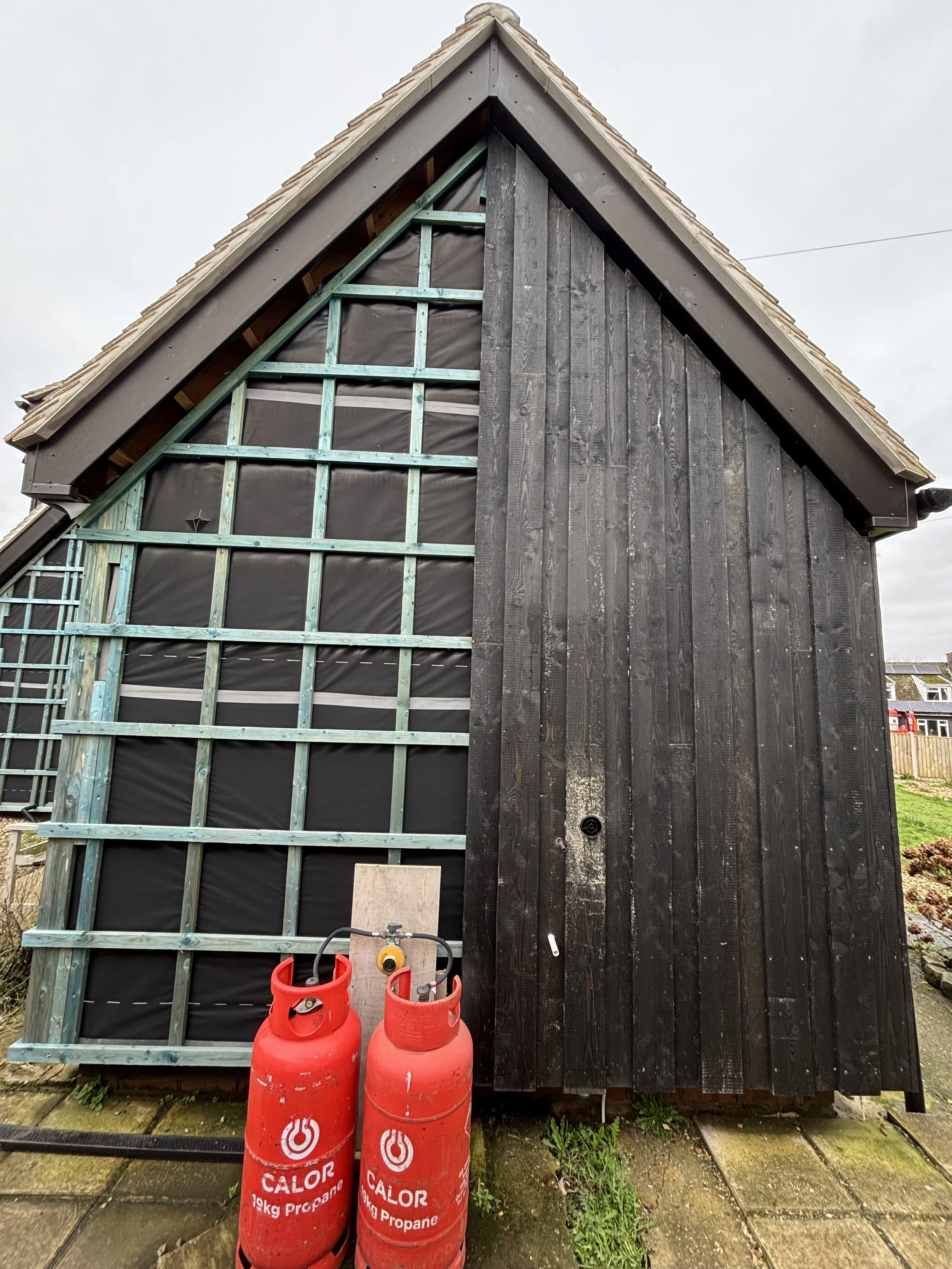 The photo shows the front of a building under construction or renovation, with some wood framework and black screen installed on the upper part. There are two red propane gas cylinders labeled 'CALOR 19kg Propane' in front of the building, and various gardening or construction tools nearby. The building has a sloped roof and vertical wooden siding.