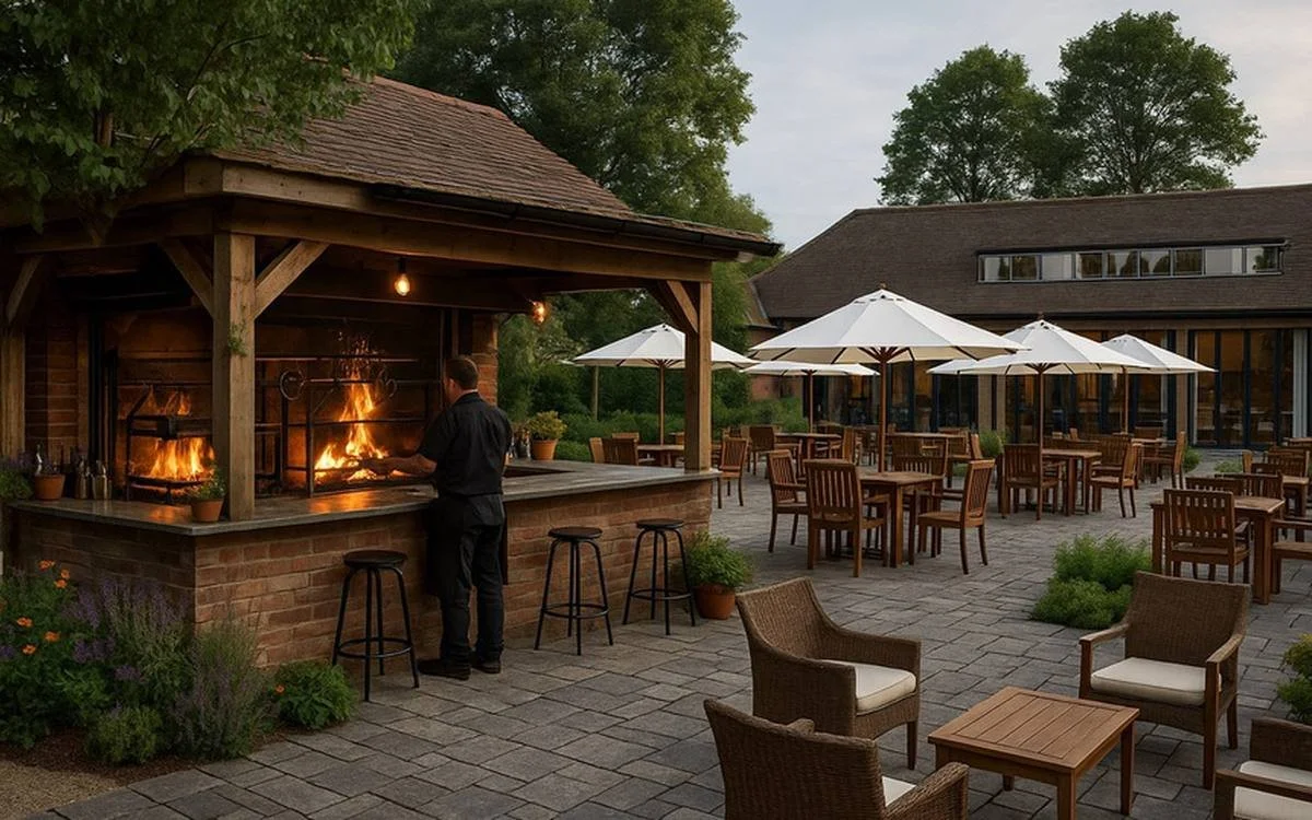 Outdoor restaurant patio with a brick barbecue grill, a chef tending to a fire, wooden tables with white umbrellas, and cushioned chairs surrounded by greenery.