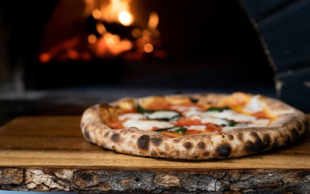 A pizza being baked inside a wood-fired oven with flames visible in the background.