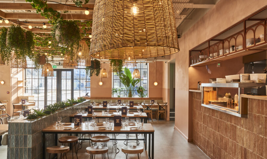 Interior of a modern restaurant with hanging wicker pendant lights, green plants, a long communal table, and a counter with open kitchen area.