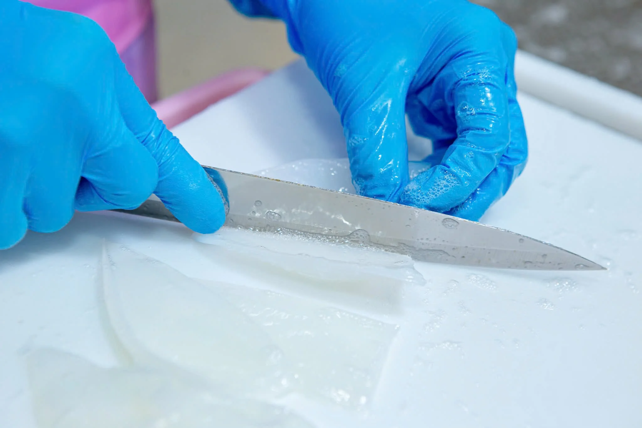 Close-up of hands wearing blue gloves scraping a white surface with a metal tool, possibly for cleaning or preparation.