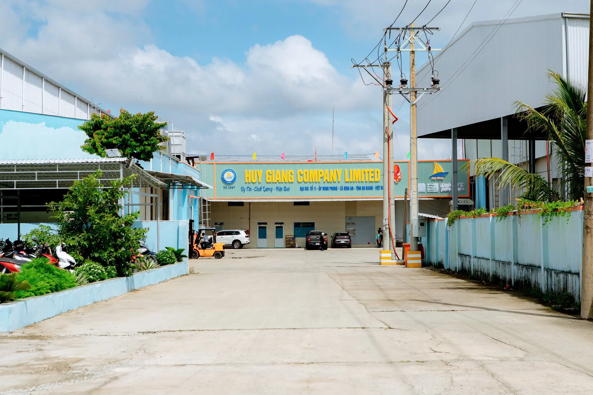 Industrial building with a sign reading 'Huy Giang Company Limited,' parked cars, trees, and power lines under cloudy sky.