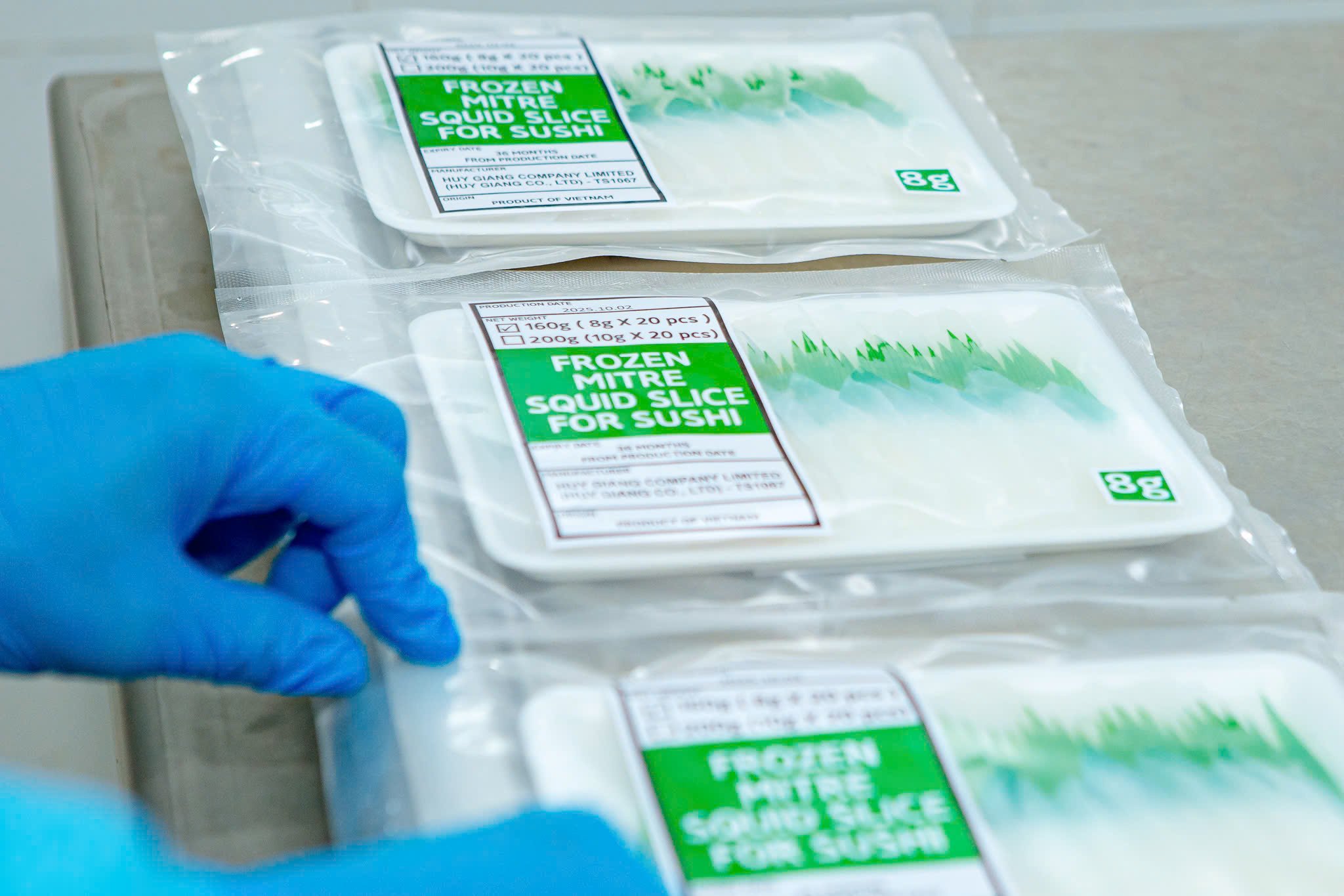 Close-up of packaged frozen squid slices for sushi on a stainless steel surface, with a gloved hand in the foreground.