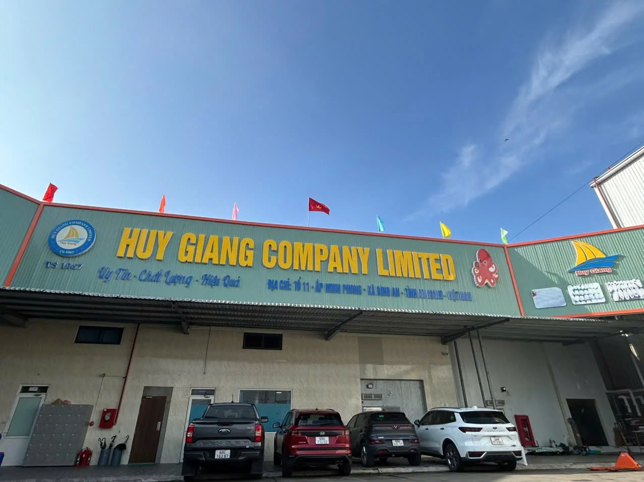 Sign for Huy Giang Company Limited with parked cars in front, flags on top, and a blue sky background.