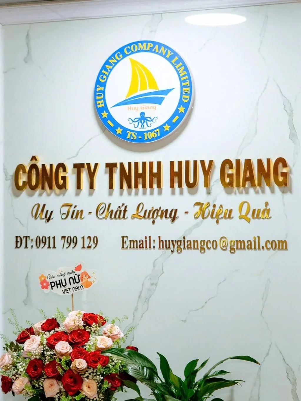Signboard and flowers at Huy Giang Company Limited, with company logo featuring sailboat and octopus, and text in Vietnamese with contact information.