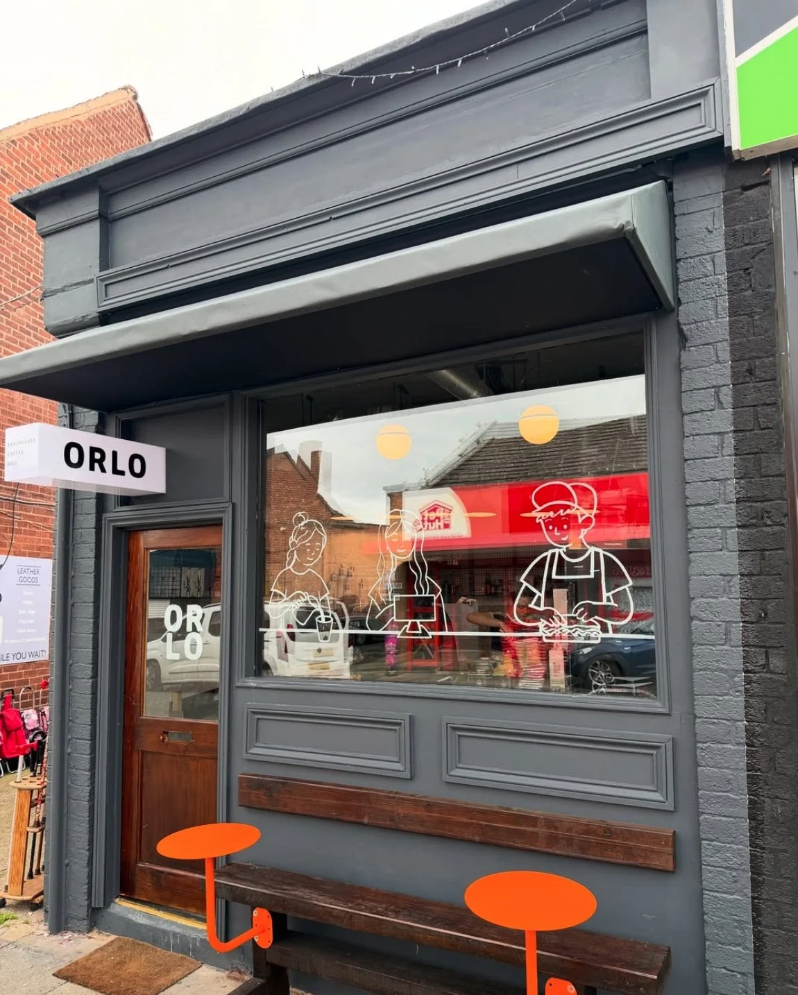 Exterior of a grey-painted storefront with a wooden door, large window with white line art illustrations of people, a red interior sign, and a sign reading 'ORLO'. There are two orange round stools outside.