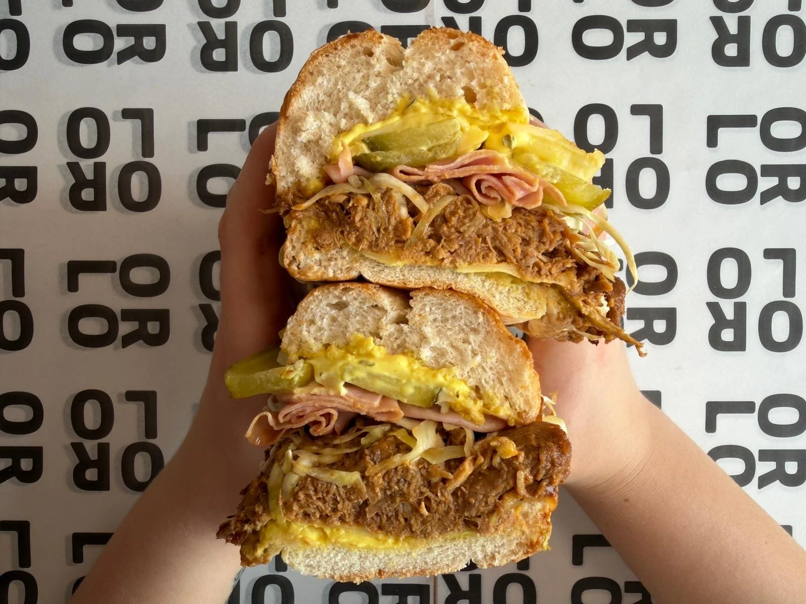 Person holding two large sandwiches filled with meat, pickles, and mustard, on a white surface with black letters.