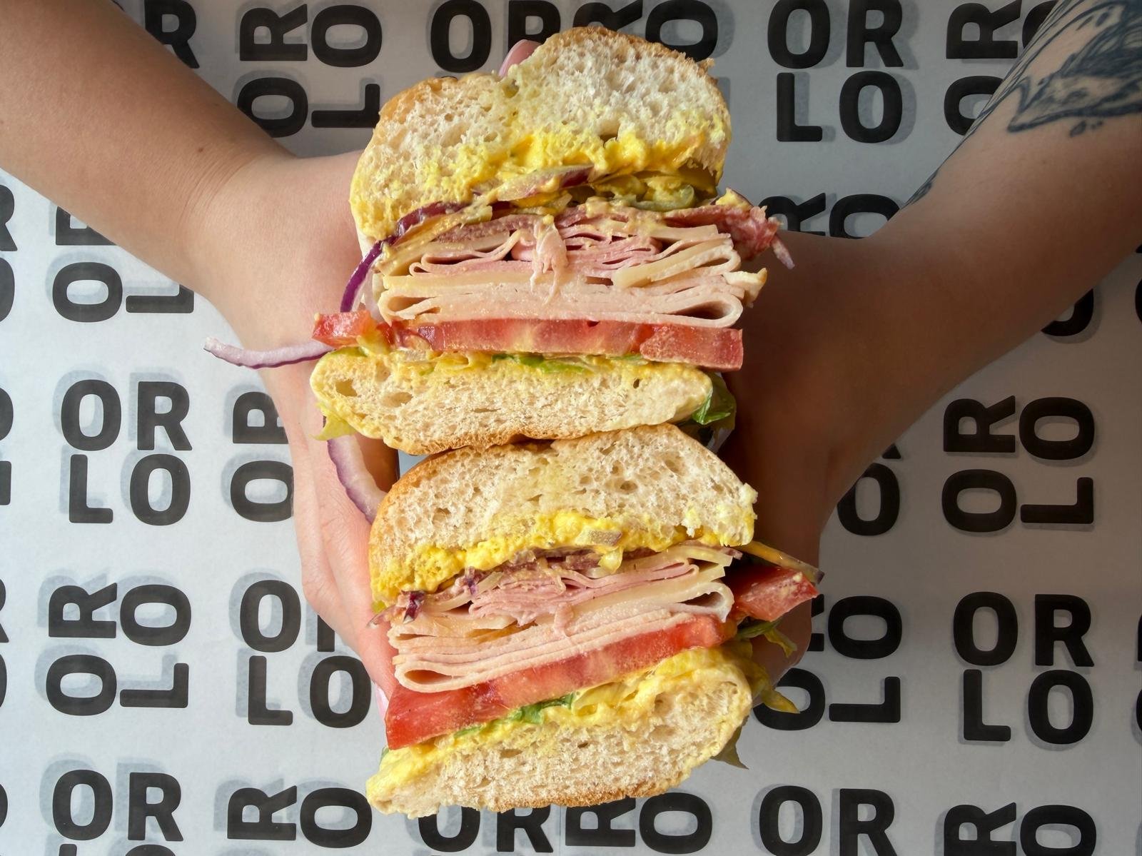 A large sandwich with multiple layers of deli meats, eggs, lettuce, tomato, and onion, held between halves of a soft bun. The sandwich appears to be from a sandwich shop, with a background featuring a design with repeating black text on white.