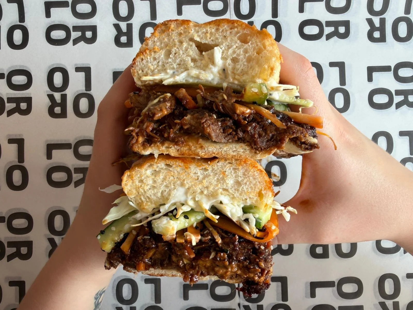Close-up view of a hand holding a stacked sandwich with various fillings, including beef, vegetables, and sauce, on a white and black patterned table.