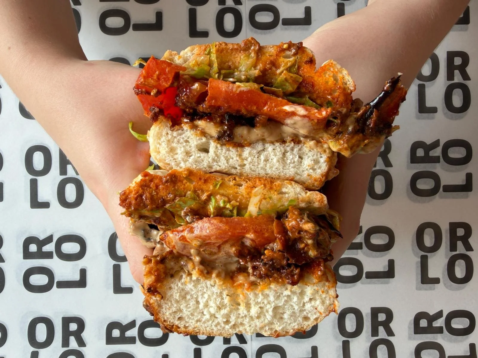 Half of a pizza slice sandwich held in hand with tomato, cheese, green pepper, and meat topping, on a paper with ORLO' printed repeatedly.