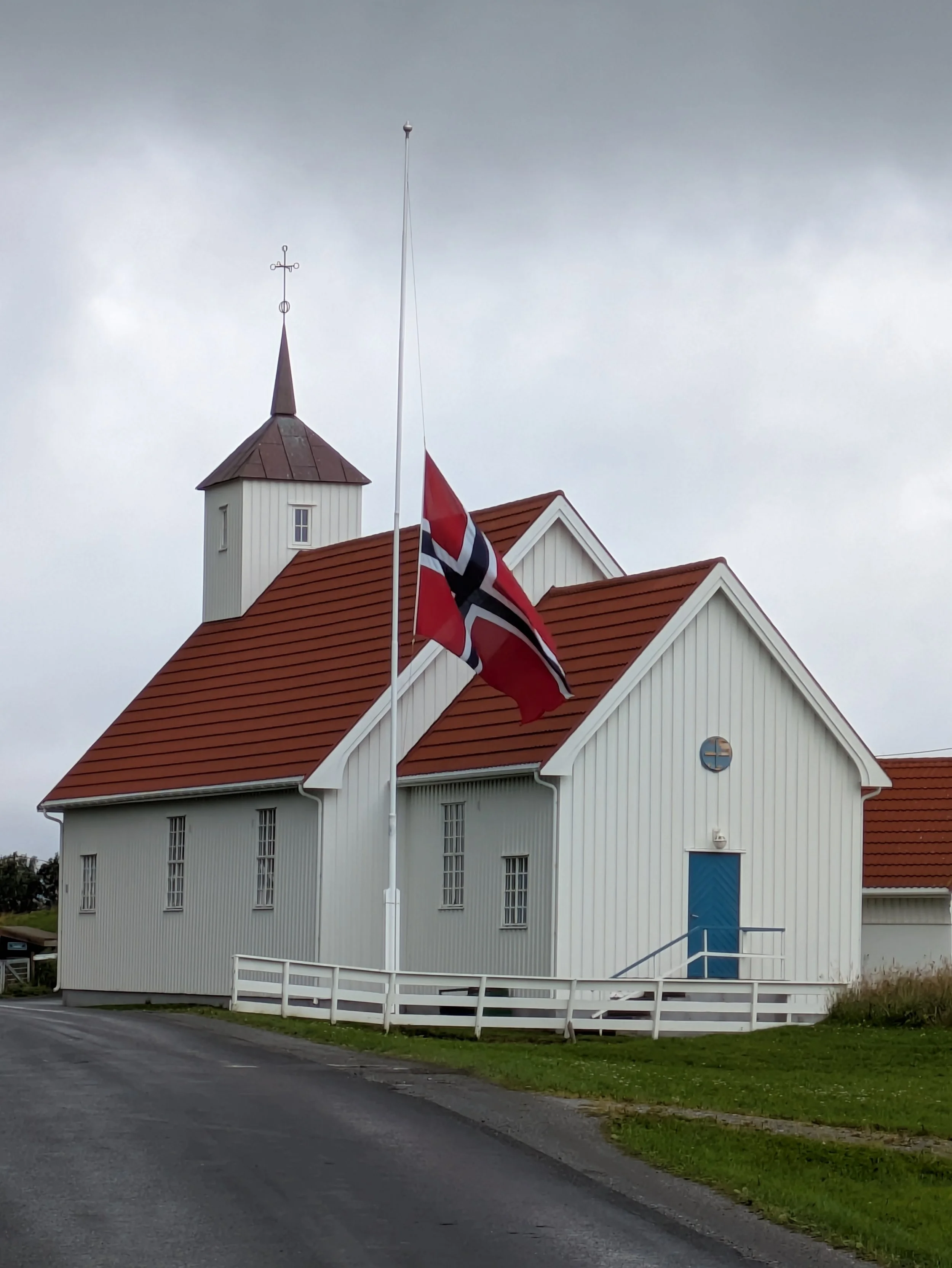 En hvit trebygning med rødt tak og blå dør, omgitt av en hvit rekkverk, med et værhus og flaggstang foran, under en overskyet himmel.