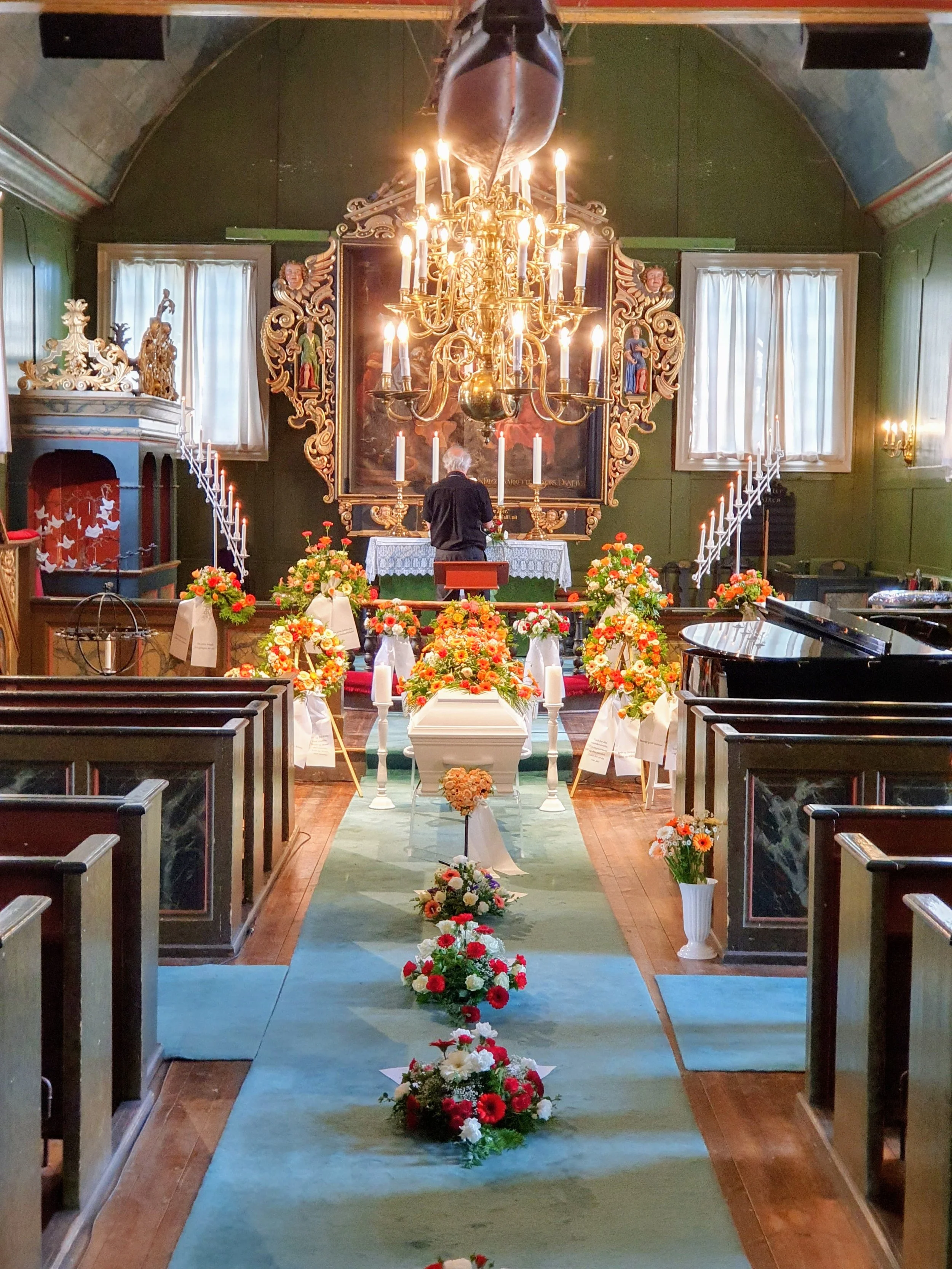 Interiør av en kirke med et alter dekorert med blomster, lysestaker og et kors. Sentrisert foran alteret er en person som står dukket opp, omgitt av flere flower arrangements og en grønn løper. Veggene er grønne med dører med hvite gardiner, og store lysekroner henger fra taket.