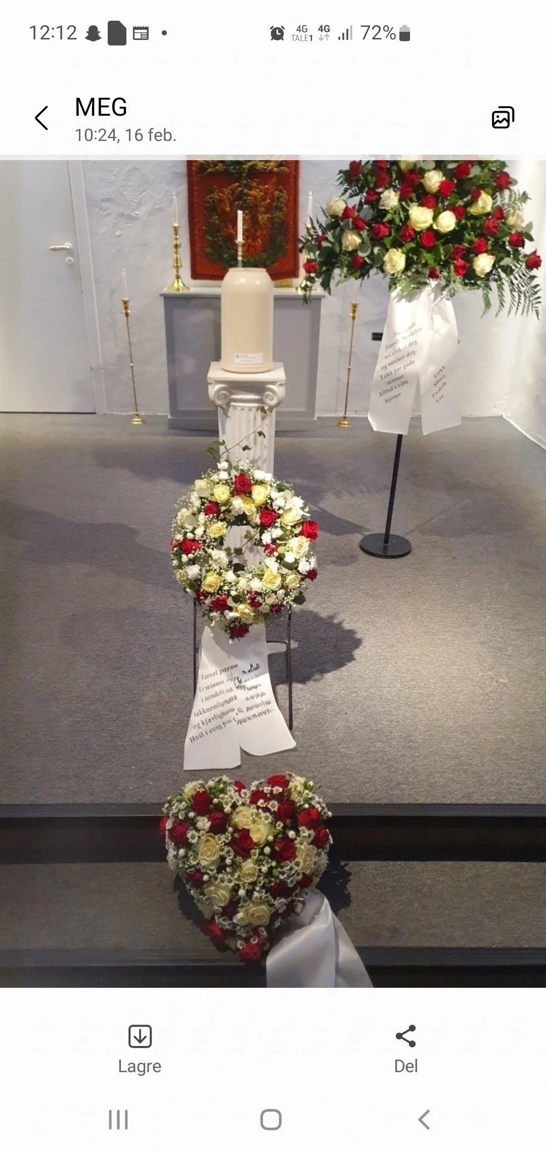 En blomsterkrets med hvite og røde roser, plassert på et stativ. I bakgrunnen er det en lys figur, en varmepumpe, og noen lysestaker. Et bilde henger på veggen bak.