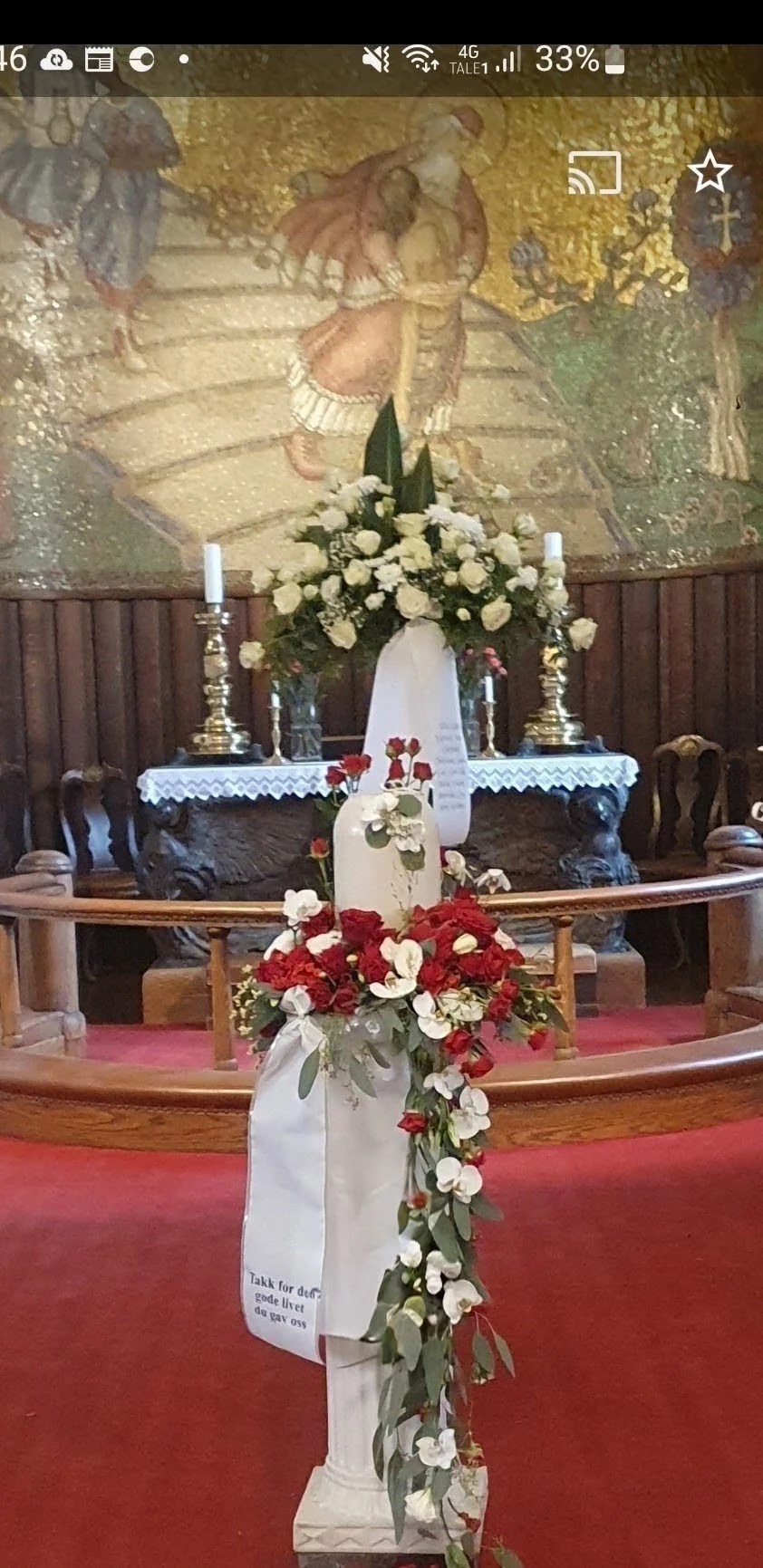 Et alter med en stor blomsteroppsats av hvite roser, omgitt av små lys og andre blomster, i en kirke med treseter og en maleri av en religiøs scene i bakgrunnen.