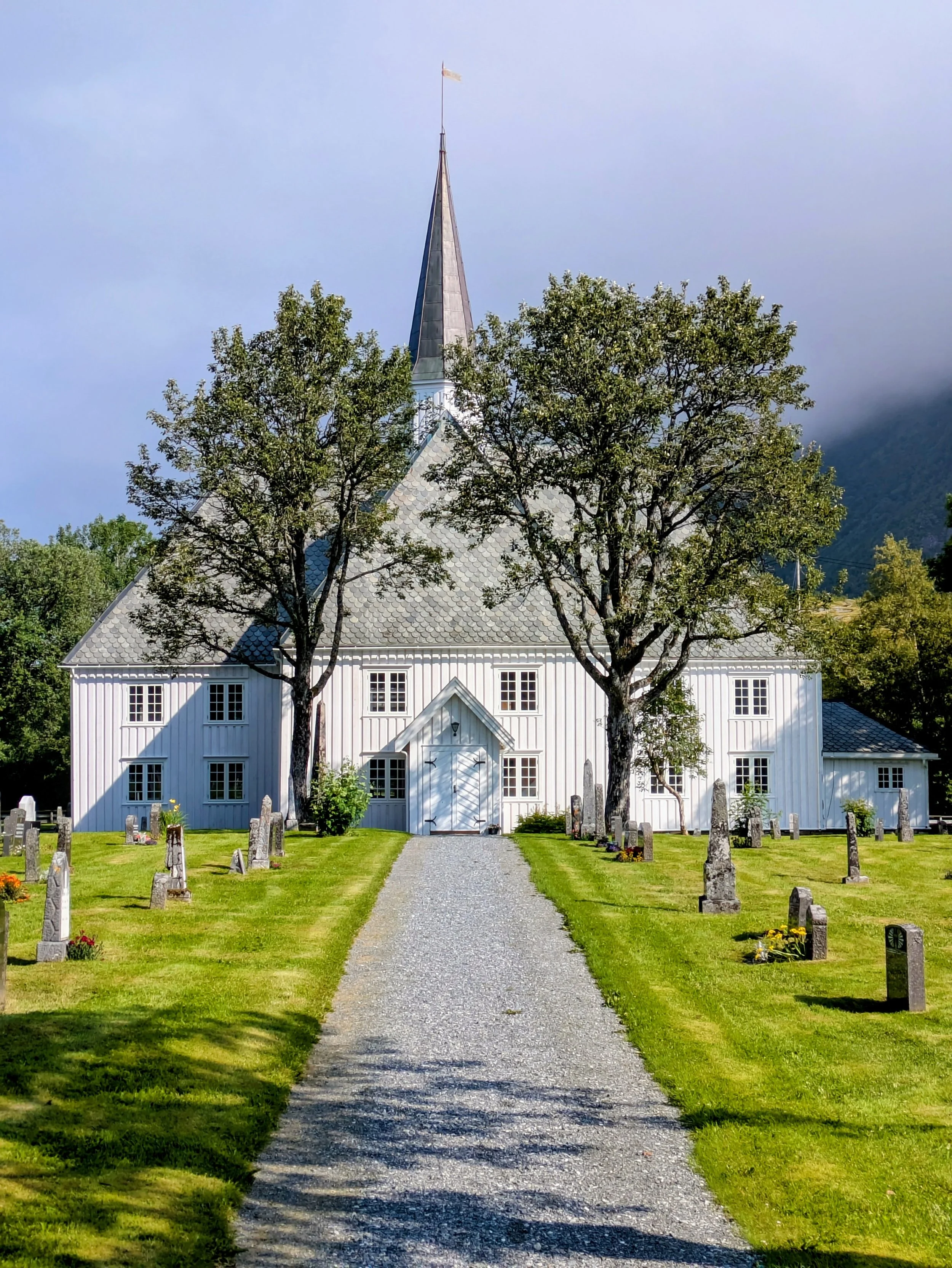 En hvit kirke med spiss tak, omgitt av to store trær, med en grusvei som fører opp til inngangen, omgitt av en godt vedlikeholdt gressplen med gravlund på begge sider.