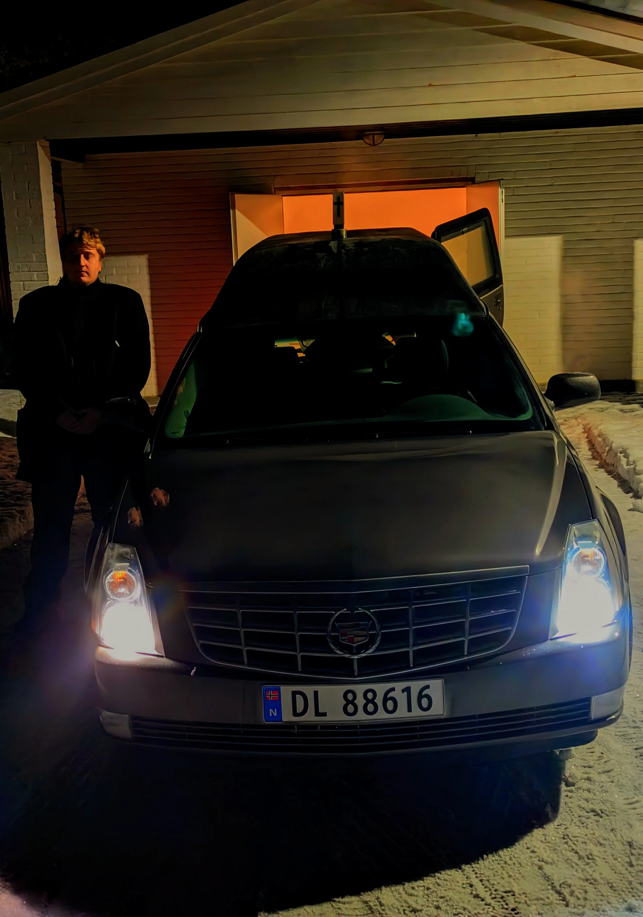 En sort Cadillac med norske registreringsskilt DL 88616, parkert utenfor en garasje under mørket. En person med mørk jakke står ved førersetet, garasjedøren er delvis åpen og det er lys inni garasjen.