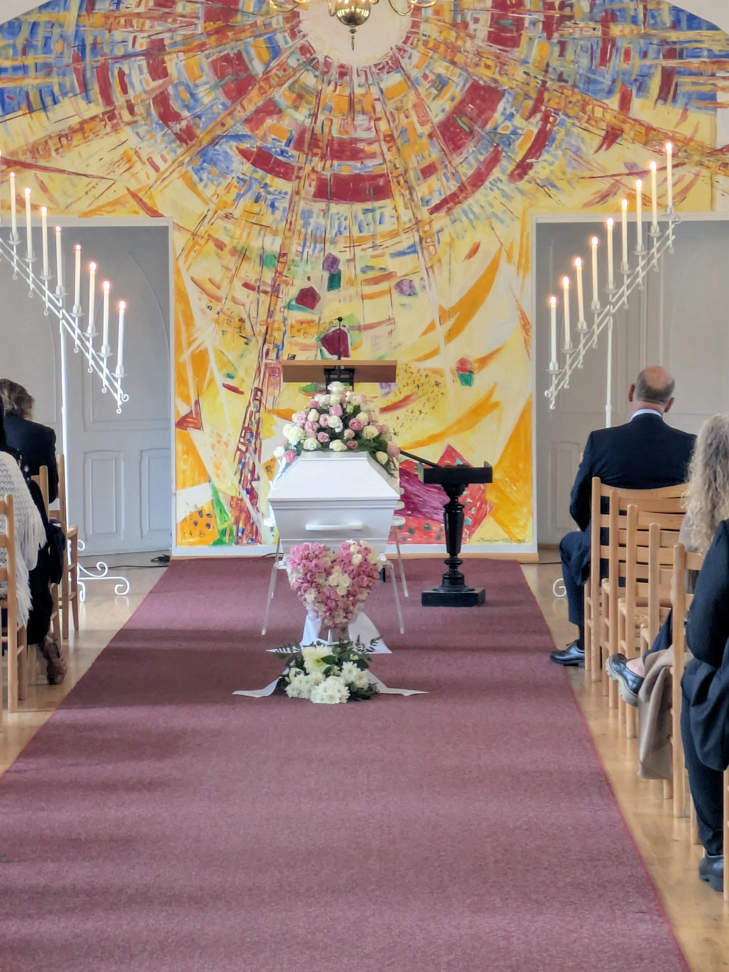 Innendørs bryllupssted med fargerikt, abstrakt takmaleri og lanterner langs midtgangen. Det er blomsterdekorasjoner foran alteret, der noen personer er til stede.