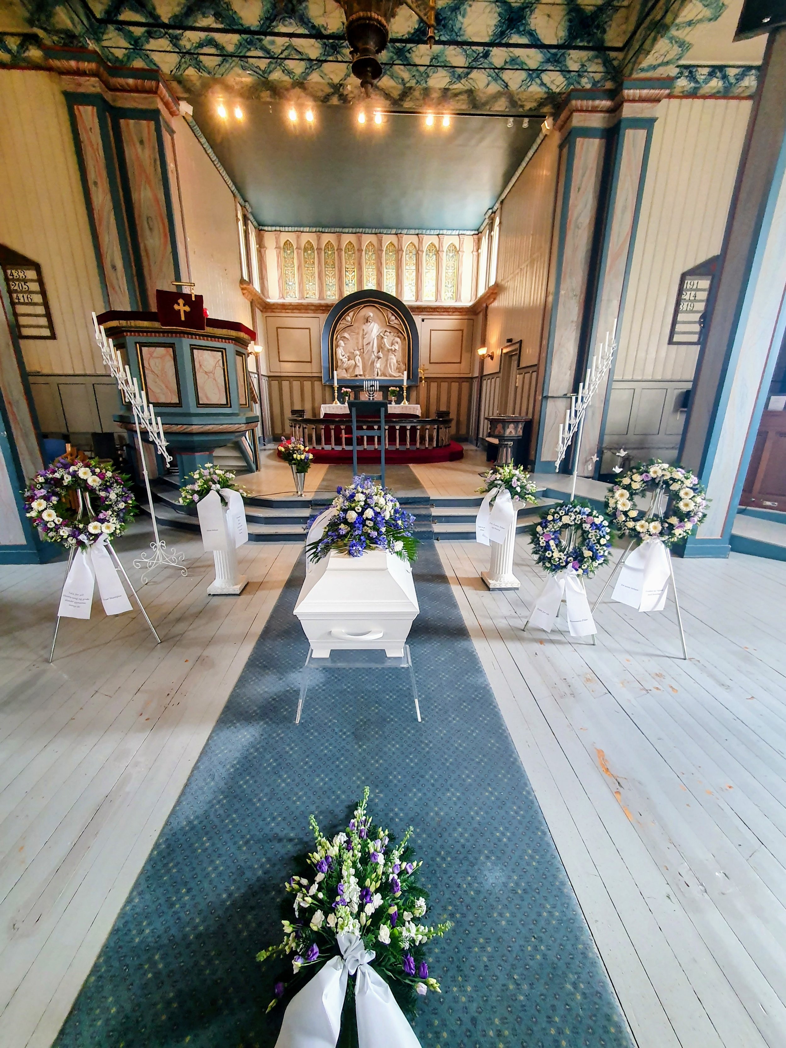 Interiør av en kirke med alteret i bakgrunnen, pyntet med blomster og blomsterarrangementer foran alteret. Det er blomsterkranser plassert på lodder langs midtgangen, og det er et blått teppe som går nedover midtgangen.
