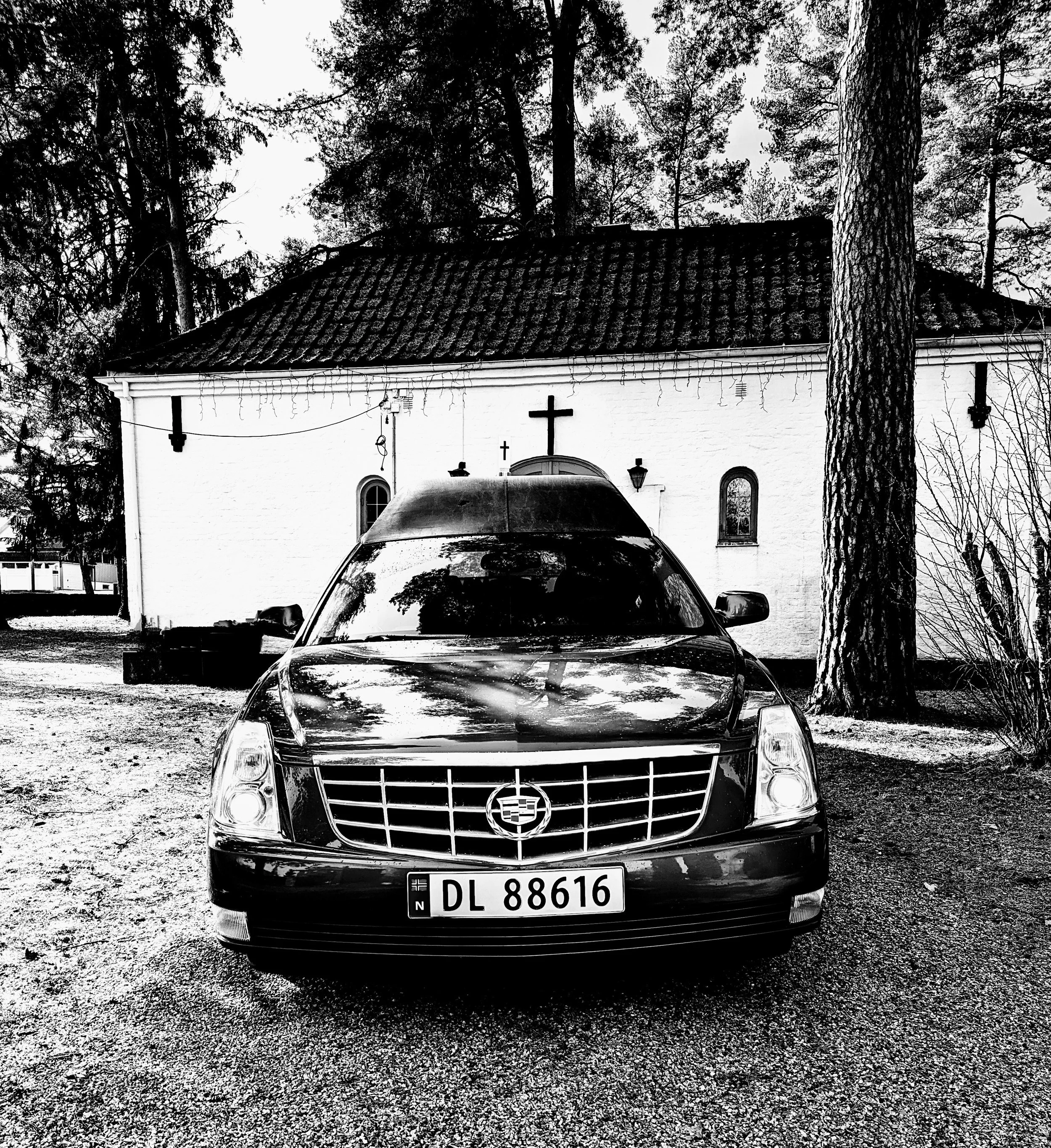 Sort og hvit bildeserie av en bil med plastdeksel foran og en kirke med flere vinduer og en kors på taket, omgitt av trær.