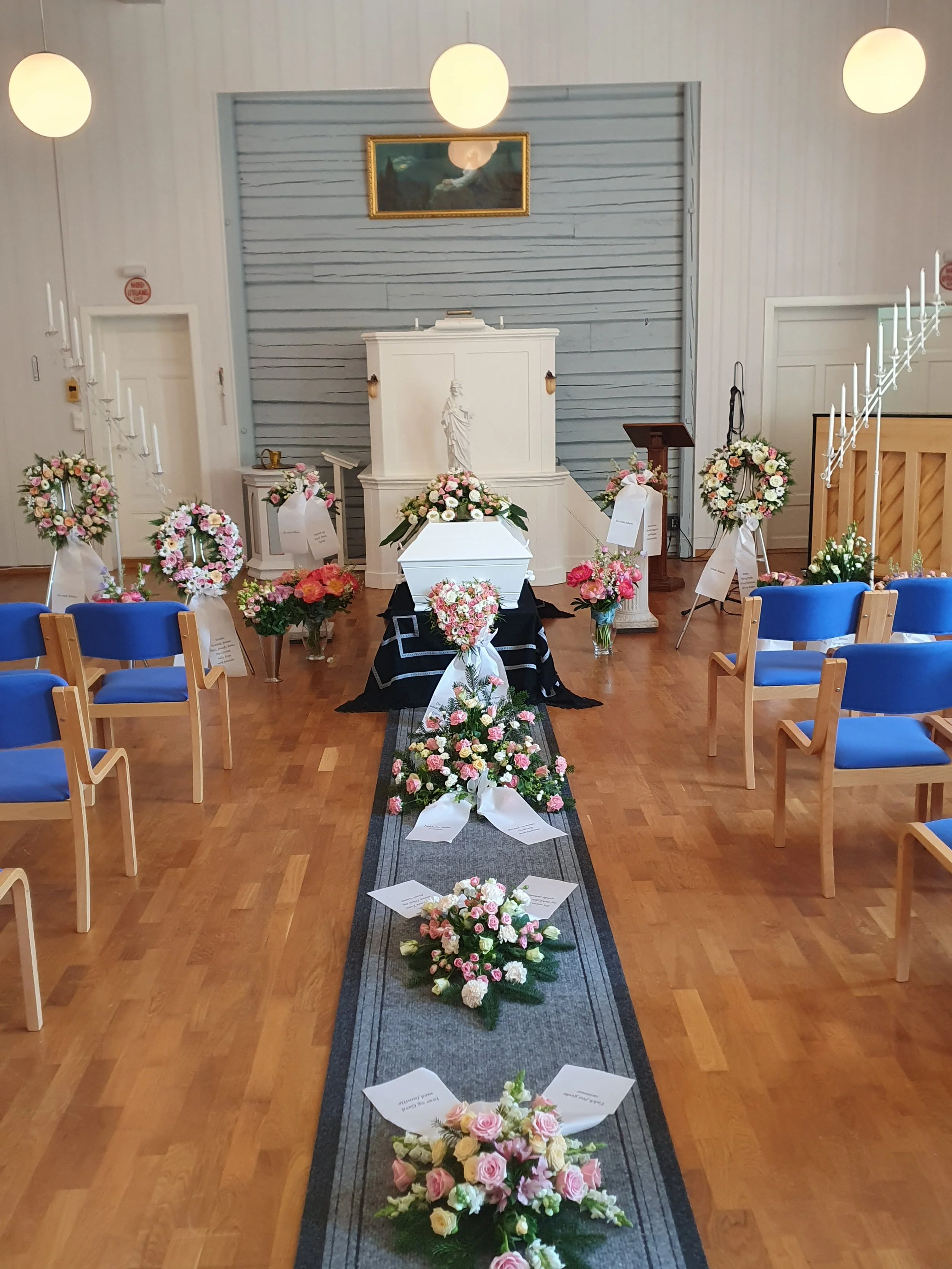 Et kirkerom pyntet for en begravelse med blomsterarrangementer langs en midtgang og et åpent kiste med blomster derpå. Det er pulter og stoler på sidene, og en altar foran.