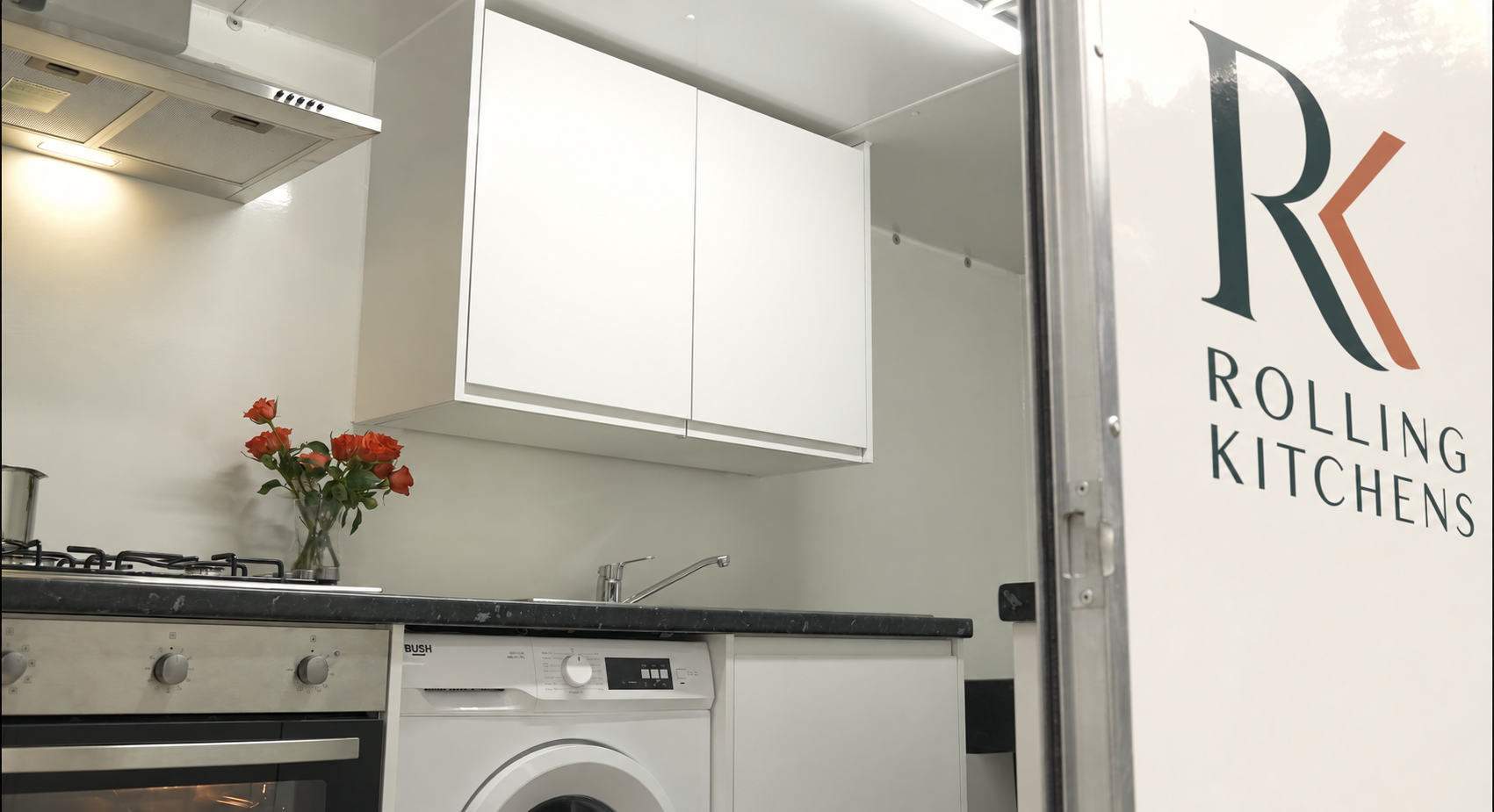 Small kitchen with white cabinets, a stove with a pot, a sink, a washing machine, a vase with red-orange roses, and a sign reading 'Rolling Kitchens' on the side of the truck.
