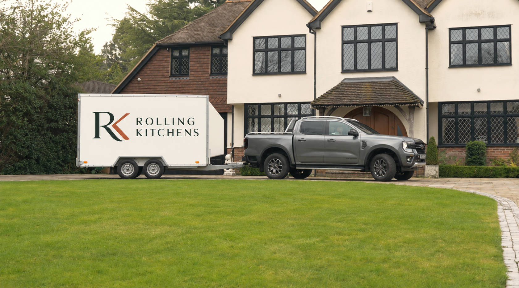A silver pickup truck with a trailer attached, parked in front of a large two-story house with a white exterior and black window frames. The trailer has the logo and text 'Rolling Kitchens' on its side.