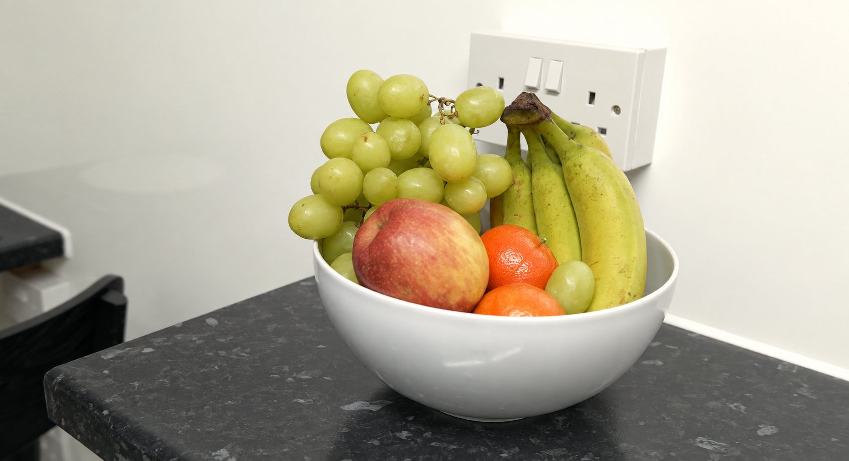 A white bowl filled with green grapes, a red apple, two oranges, and bananas sits on a black countertop near a white electrical socket.