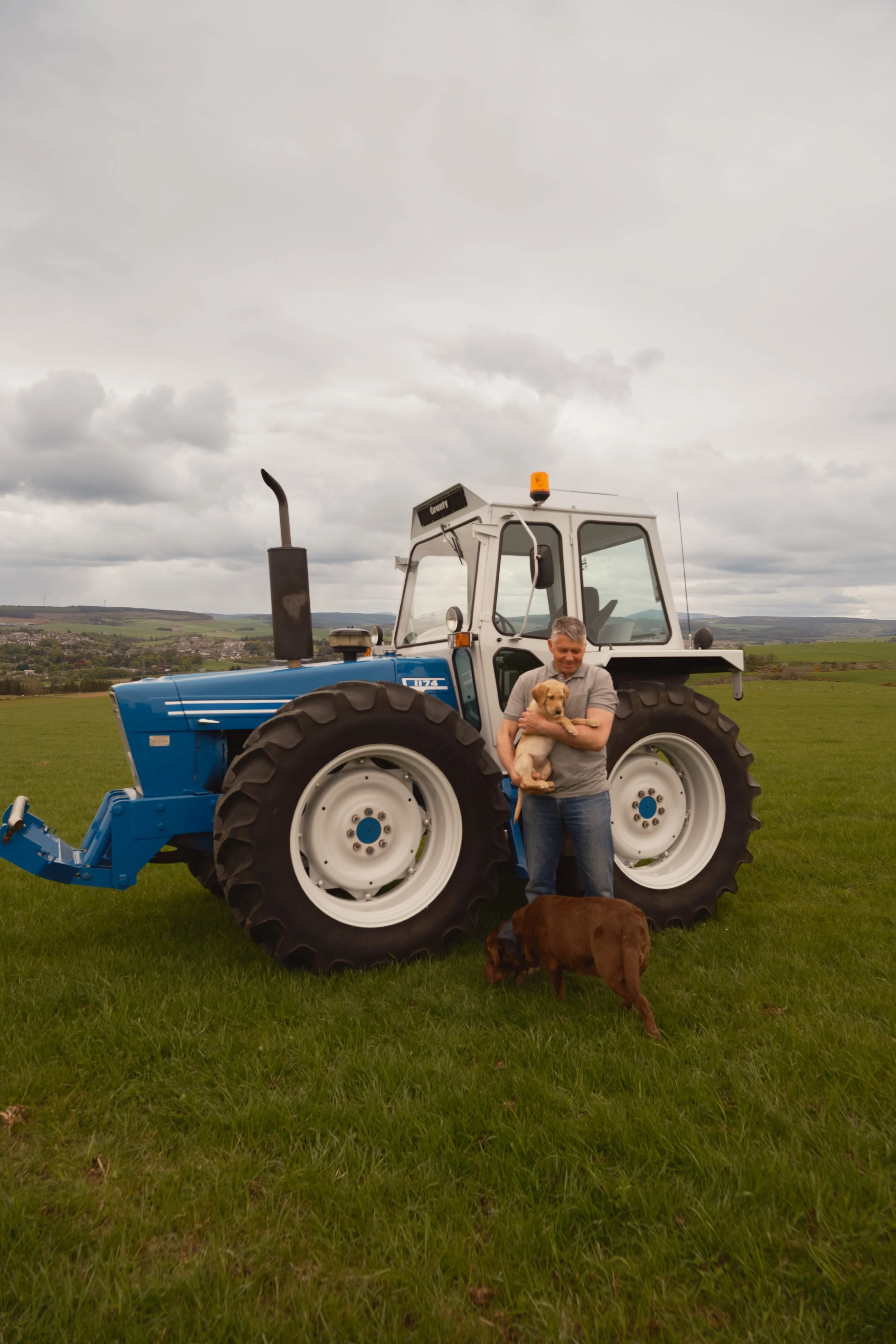 A man holding a yellow Labrador puppy standing in front of a blue tractor in a grassy field with a cloudy sky and rolling hills in the background.