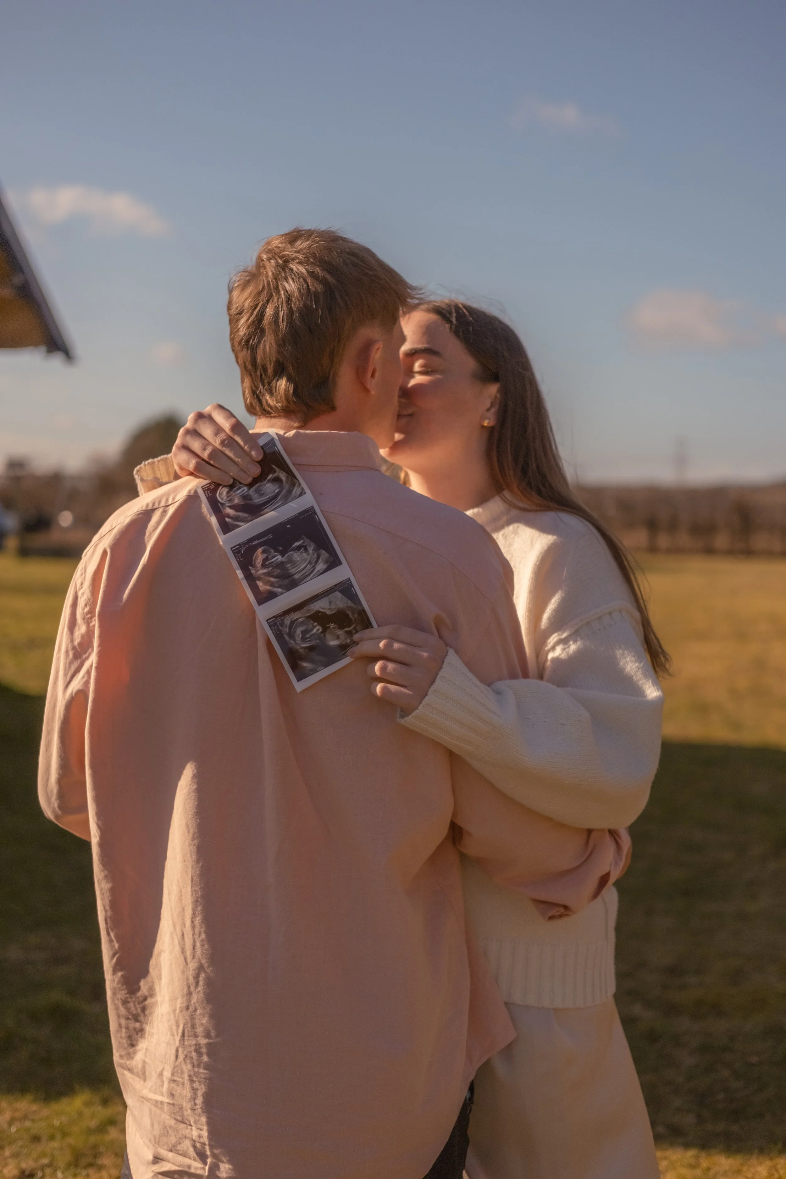 A couple sharing a kiss outdoors during the daytime, with the woman holding ultrasound images of a baby. The background features a grassy field, blue sky, and some clouds.